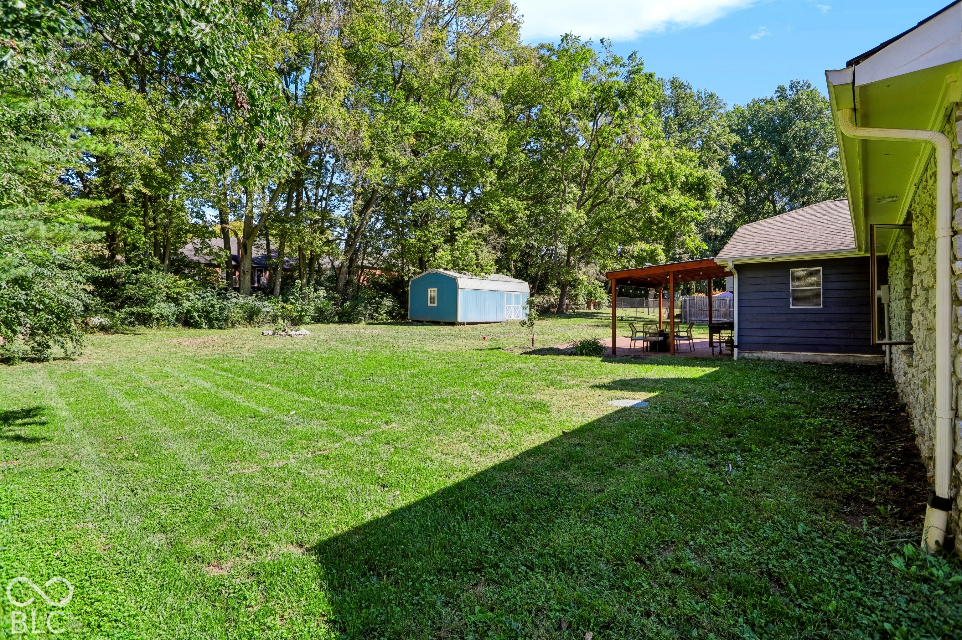 Expansive grassy yard with mature trees, a detached storage shed, and a covered patio area with a paved surface