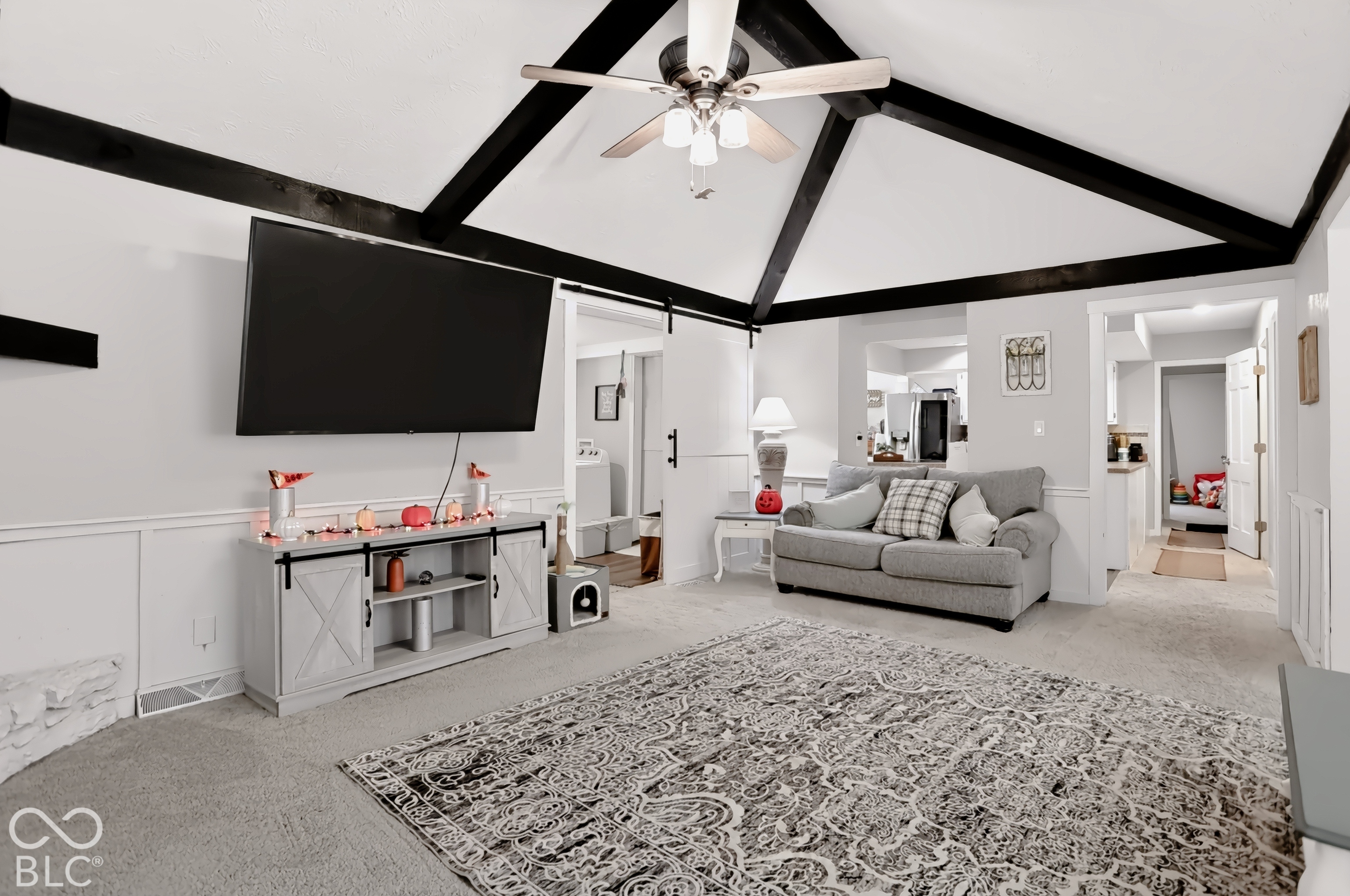 Vaulted ceiling with exposed beams, ceiling fan, white paneled wall trim, carpeted flooring, and a barn door entry