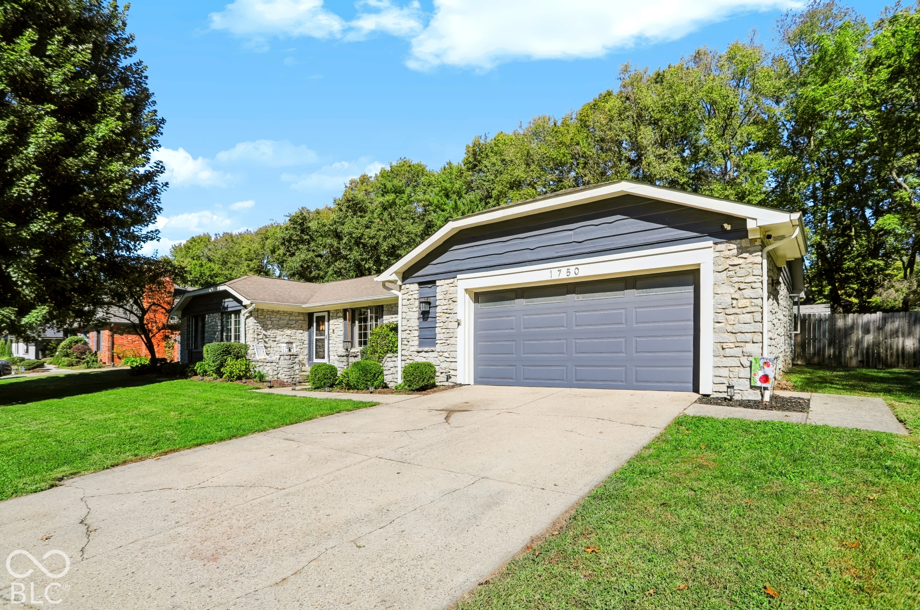Stone facade, attached garage with gray door, dark gray siding, mature trees, concrete driveway