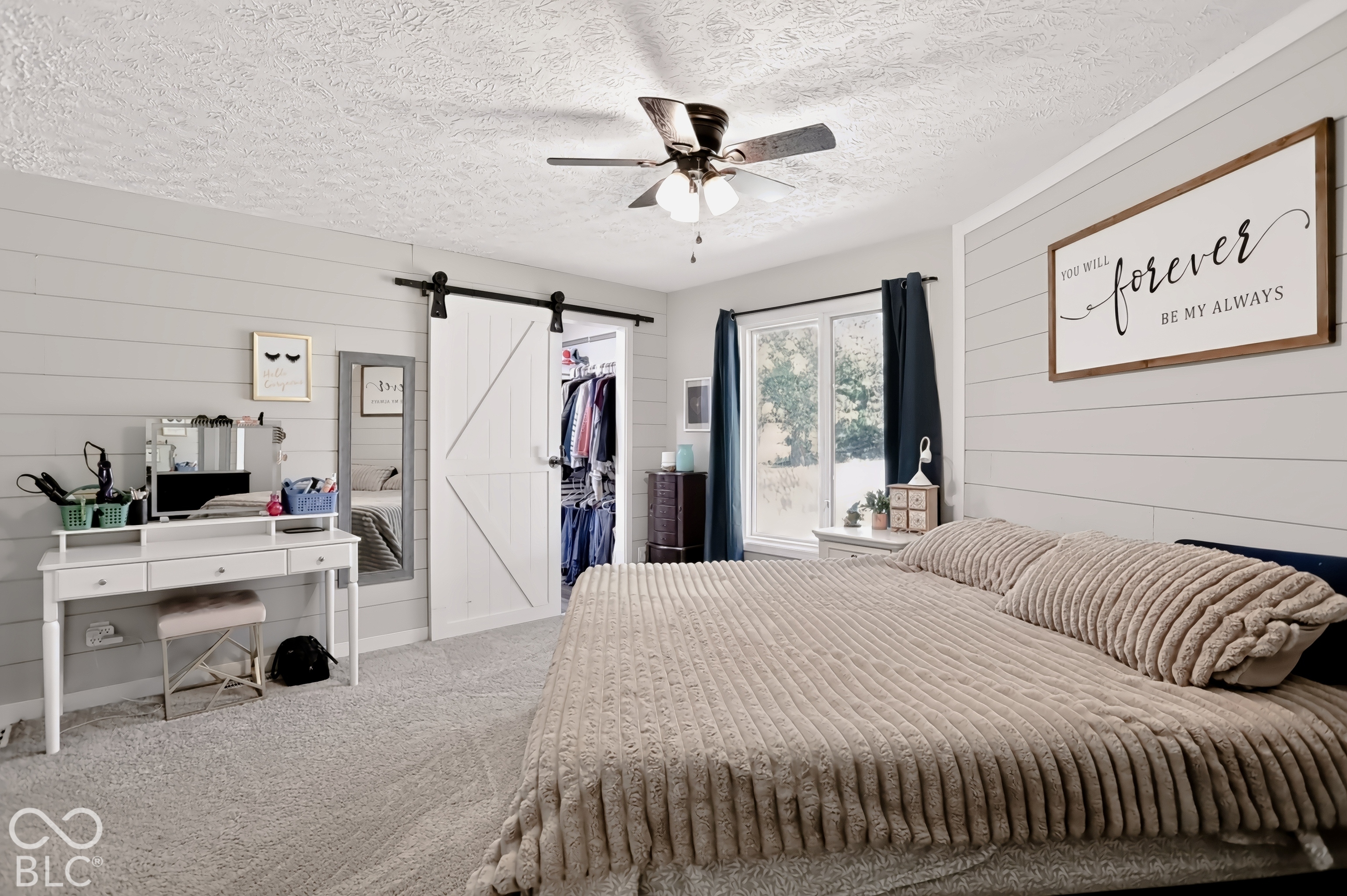 Shiplap accent wall, light-colored carpeting, ceiling fan with light fixture, large window with dark drapes, and white barn-style sliding closet doors