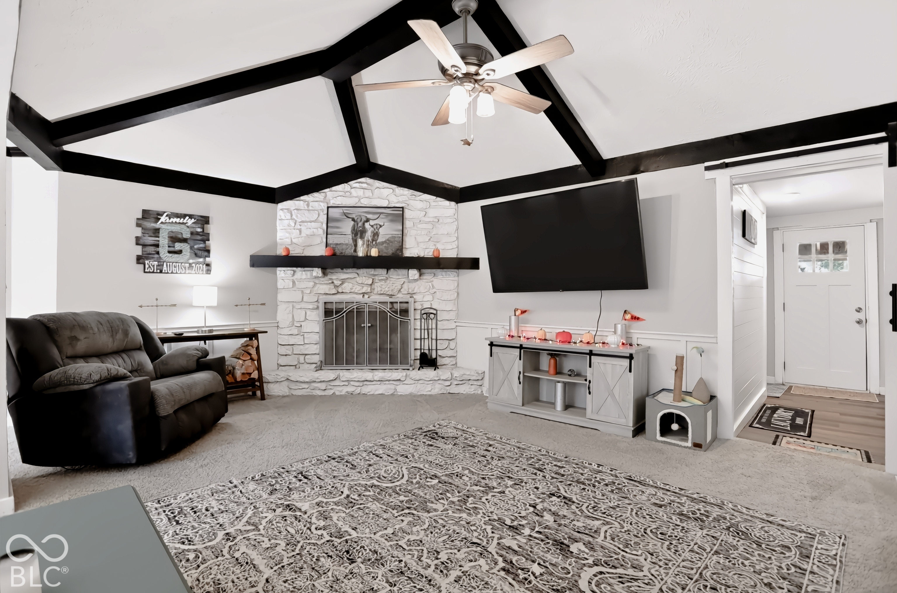 Vaulted ceiling with exposed beams, stone-clad fireplace with mantel, ceiling fan with integrated lighting, and a white paneled entry door