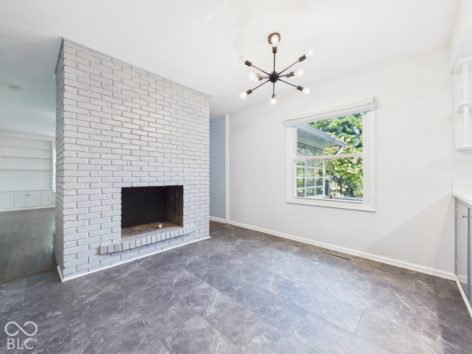 Painted brick fireplace, modern Sputnik-style chandelier, window with grid panes, built-in shelving, and grey wood-finish flooring