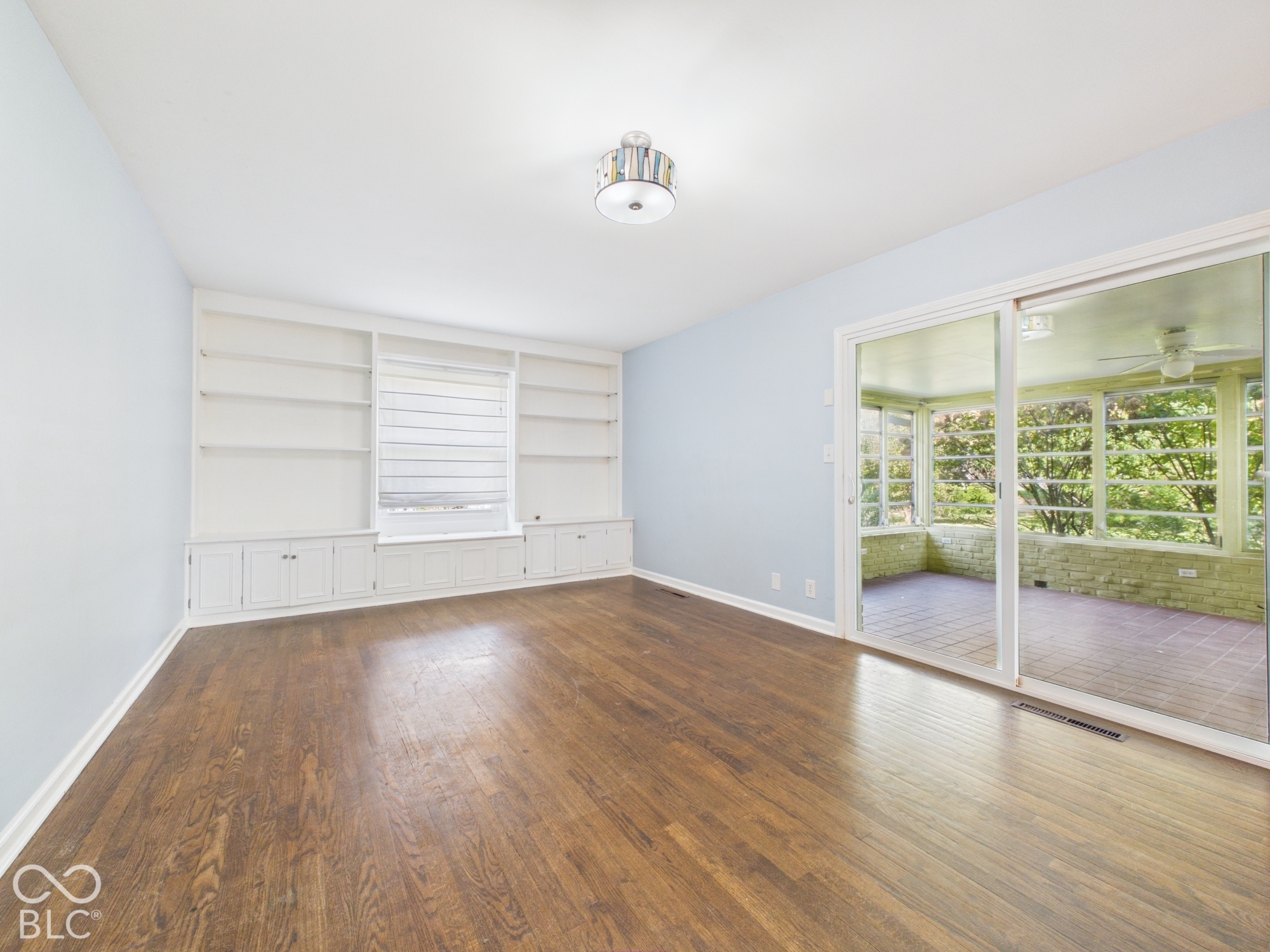 Wood-finish flooring throughout, integrated built-in shelving, window with horizontal blinds, overhead flush mount light fixture, and sliding glass doors to an enclosed sunroom with multiple windows and a ceiling fan