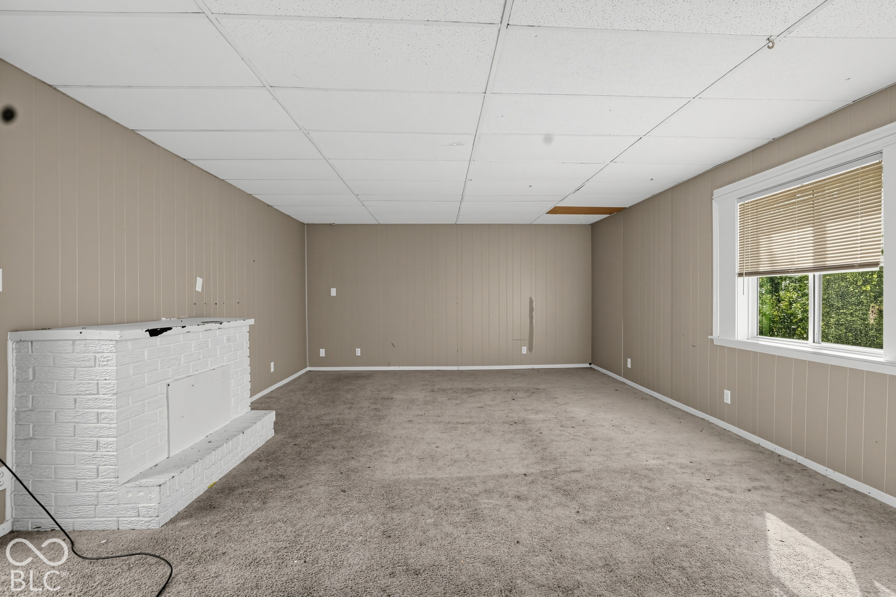 Interior space featuring vertical wood-paneled walls, a white painted brick fireplace, carpeted flooring, and a window with horizontal blinds