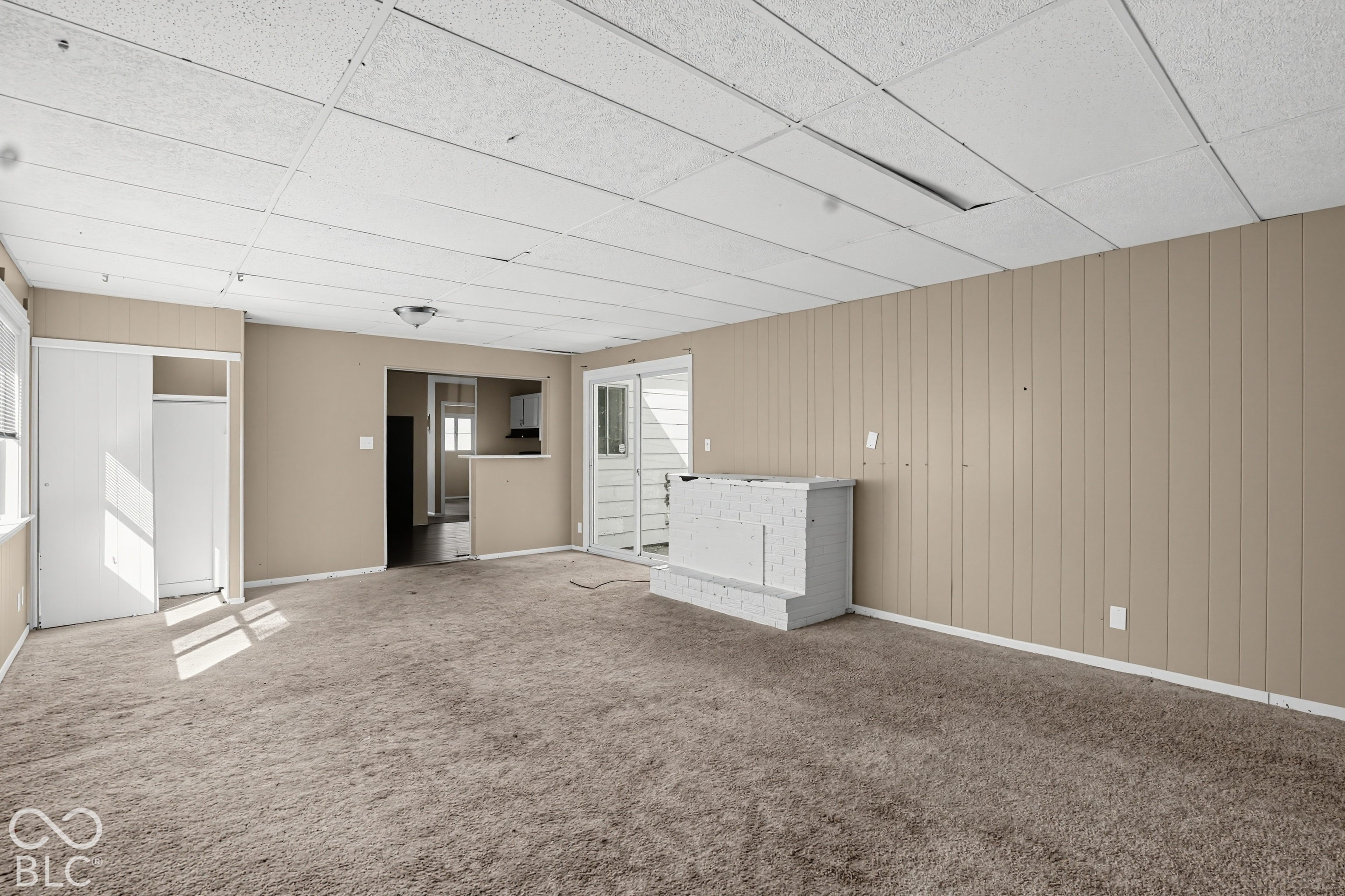 Interior space featuring carpeted flooring, paneled walls, a painted brick fireplace, a drop ceiling, and sliding glass doors