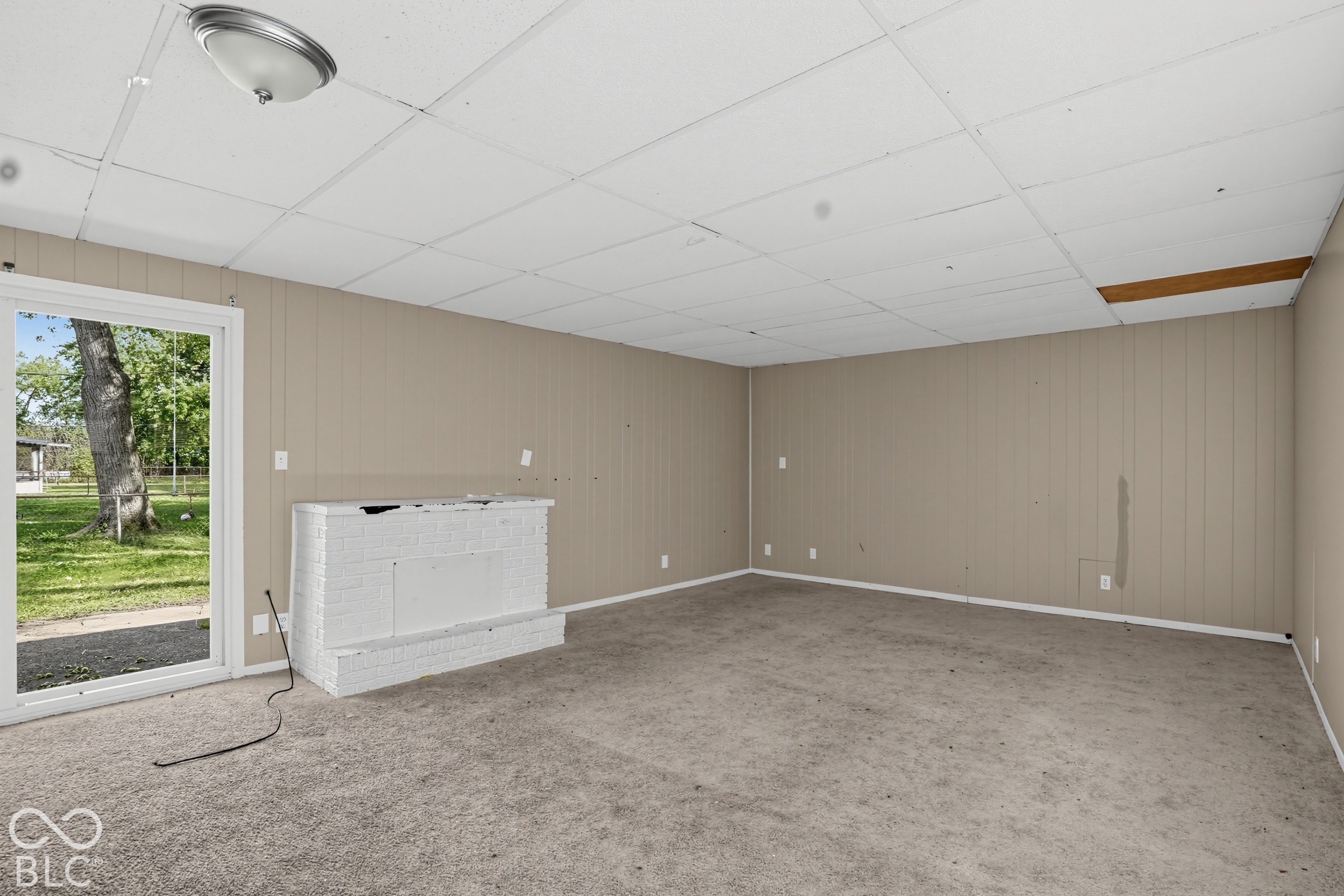 Room featuring a painted brick fireplace, paneled walls, a drop ceiling, carpet flooring, and a glass exterior door