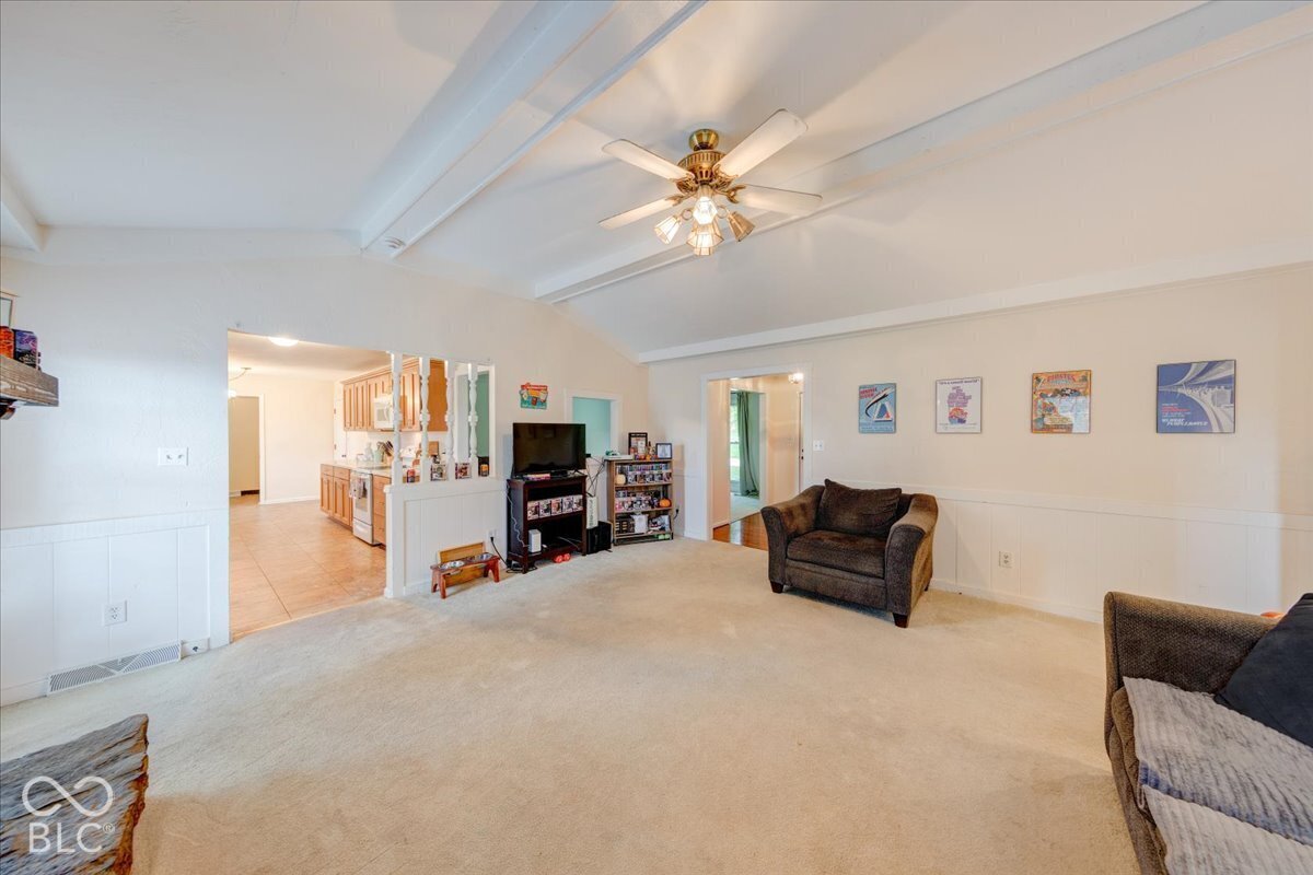 Living space featuring vaulted ceilings with exposed beams, a central ceiling fan with integrated lighting, and white wainscoting