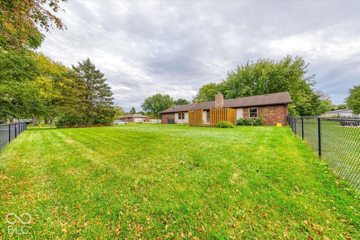 Expansive backyard featuring a level lawn, mature trees, chain-link perimeter fencing, and a brick exterior house with a stone chimney
