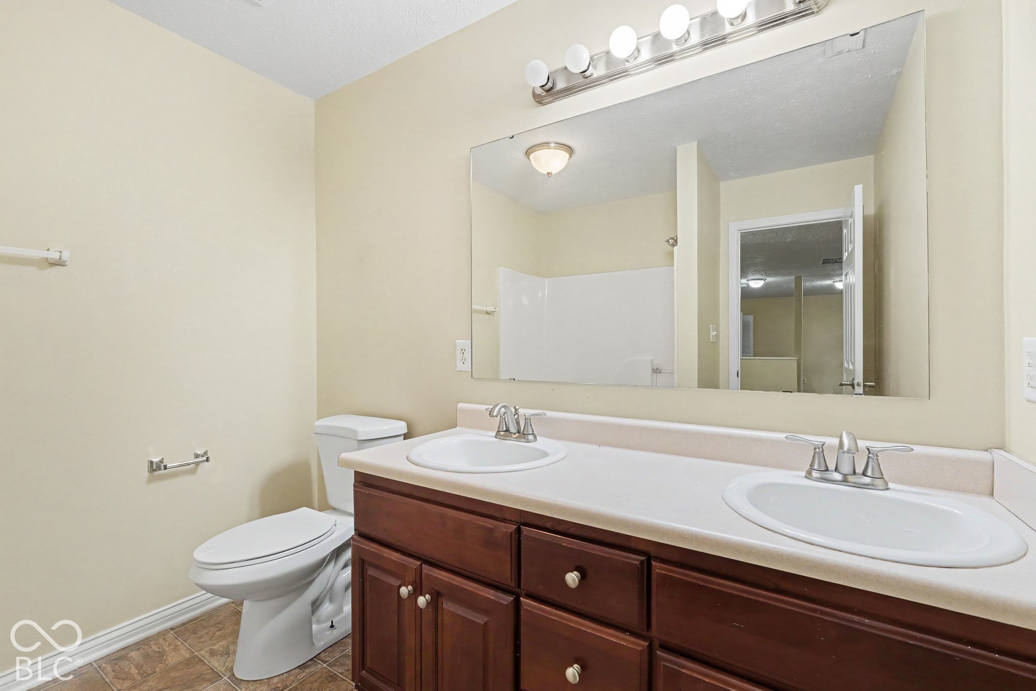 Double vanity with integrated sinks, neutral solid-surface countertop, dark wood-finish cabinetry, full-width wall mirror, and five-light vanity fixture