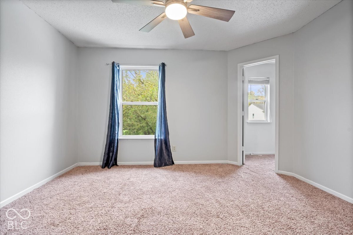 Neutral-toned interior space featuring textured wall-to-wall carpeting, a multi-blade ceiling fan with integrated lighting, and a window with natural lighting