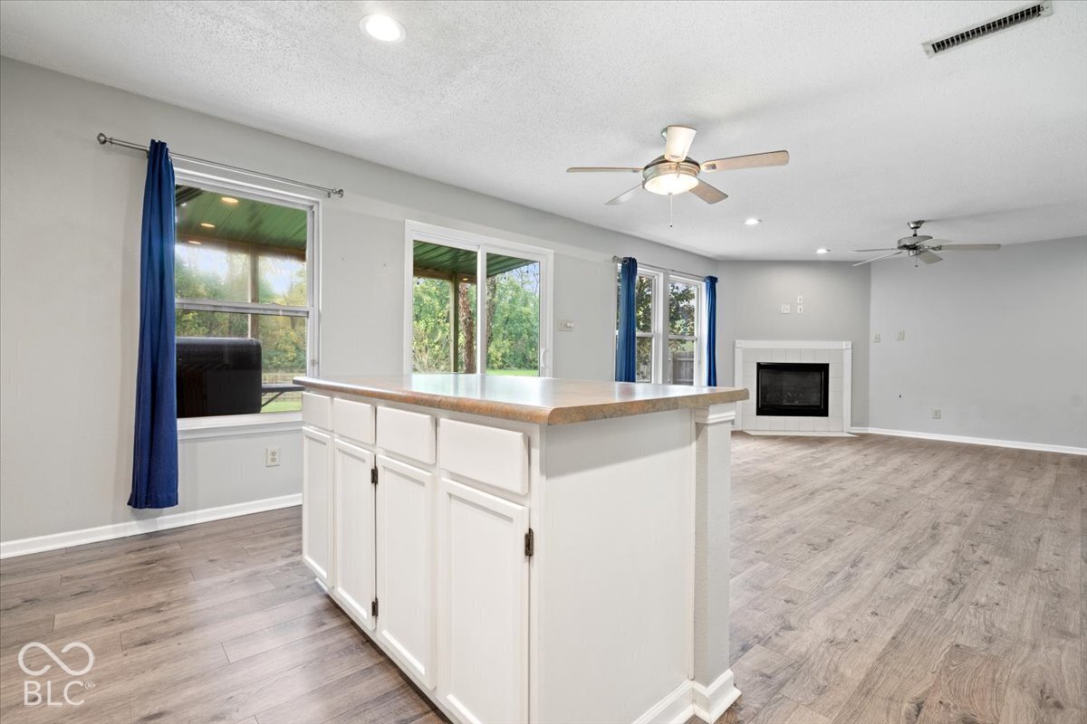 Open-concept interior featuring wood-finish flooring, a kitchen island with stone-finish countertop, dual ceiling fans, sliding glass doors, and a fireplace with a white mantle