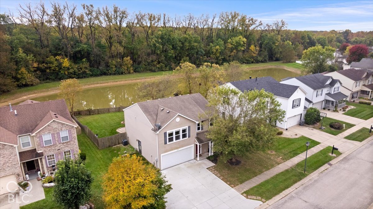 Aerial perspective featuring a two-story residence with a two-car garage, shingled roof, concrete driveway, and a fenced rear yard bordering a pond Aerial perspective featuring a two-story residence with a two-car garage, shingled roof, concrete driveway, and a fenced rear yard bordering a pond