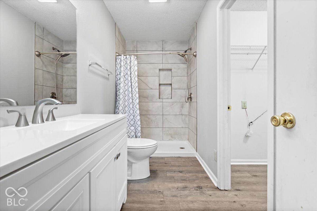 Bathroom featuring wood-finish flooring, a white vanity with a rectangular sink, large frameless mirror, and a walk-in shower with stone-style wall tile and a built-in recessed niche