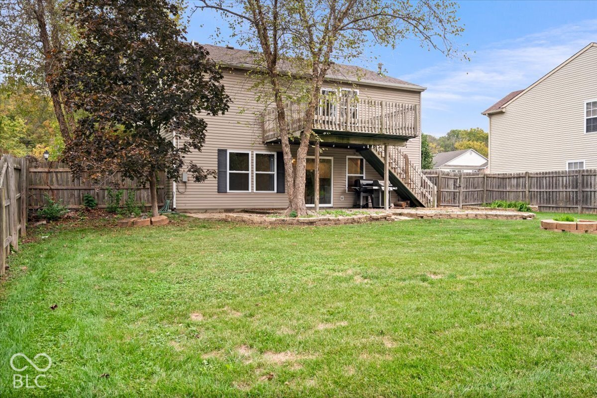Rear exterior featuring light-toned horizontal siding, a wooden elevated deck with stairs, a stone-bordered patio, and full wood perimeter fencing