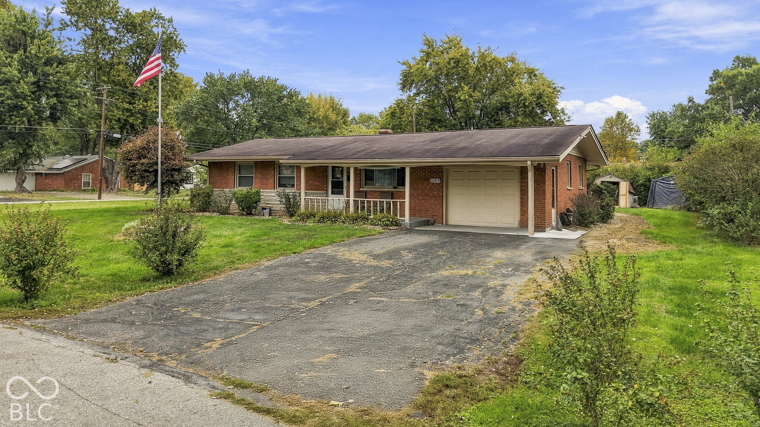 The exterior front of the ranch-style home featuring a covered front porch, an attached garage, and a concrete driveway on a landscaped lot. Flag pole in the center of the front yard.