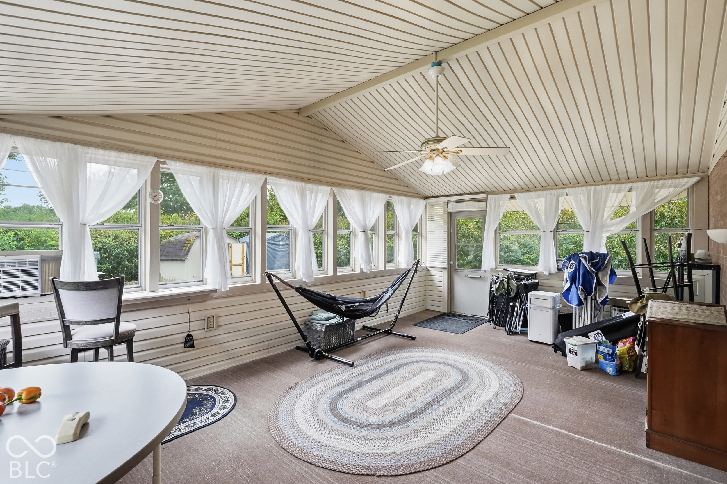 The fully enclosed sun room features a vaulted ceiling with multiple surrounding windows on all three sides, and a ceiling-mounted light fixture with a fan. The view to the back yard is calming.