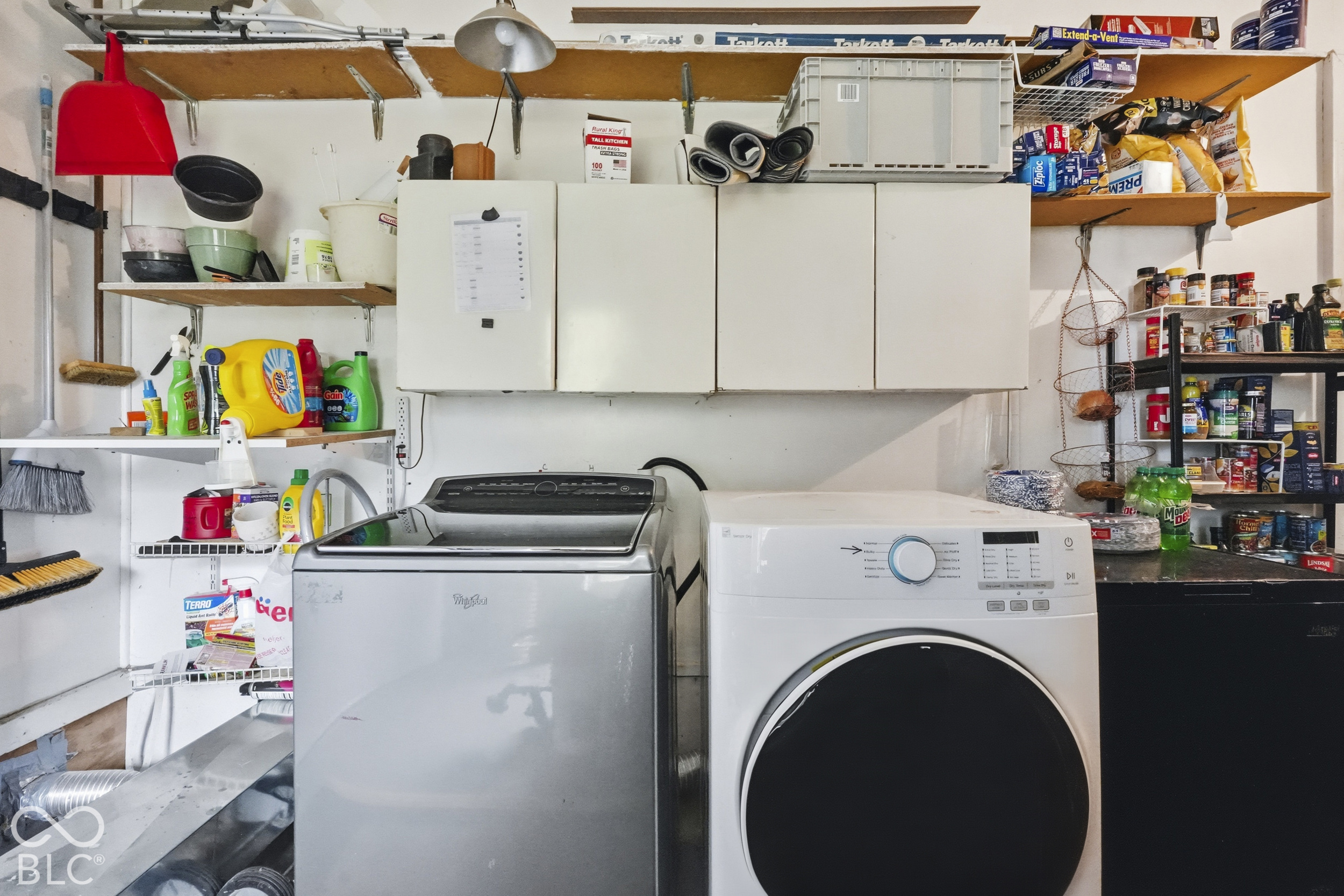 The laundry space is in the garage. There is white wall-mounted cabinetry and tiered open shelving allowing plenty of space to store supplies.