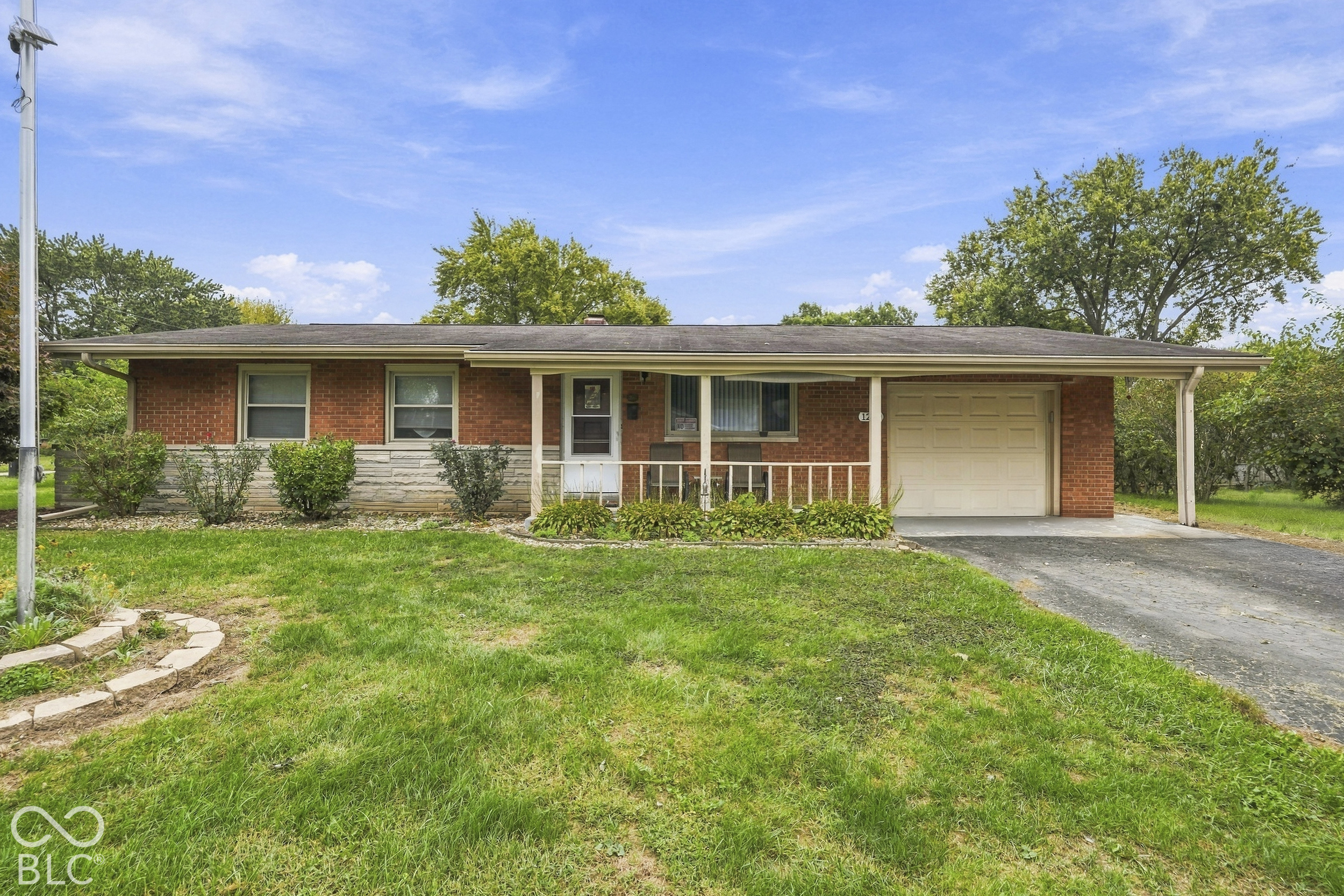 Single-story ranch exterior featuring a red brick facade with stone masonry accents, an attached one-car garage, and a covered front porch with a railing.