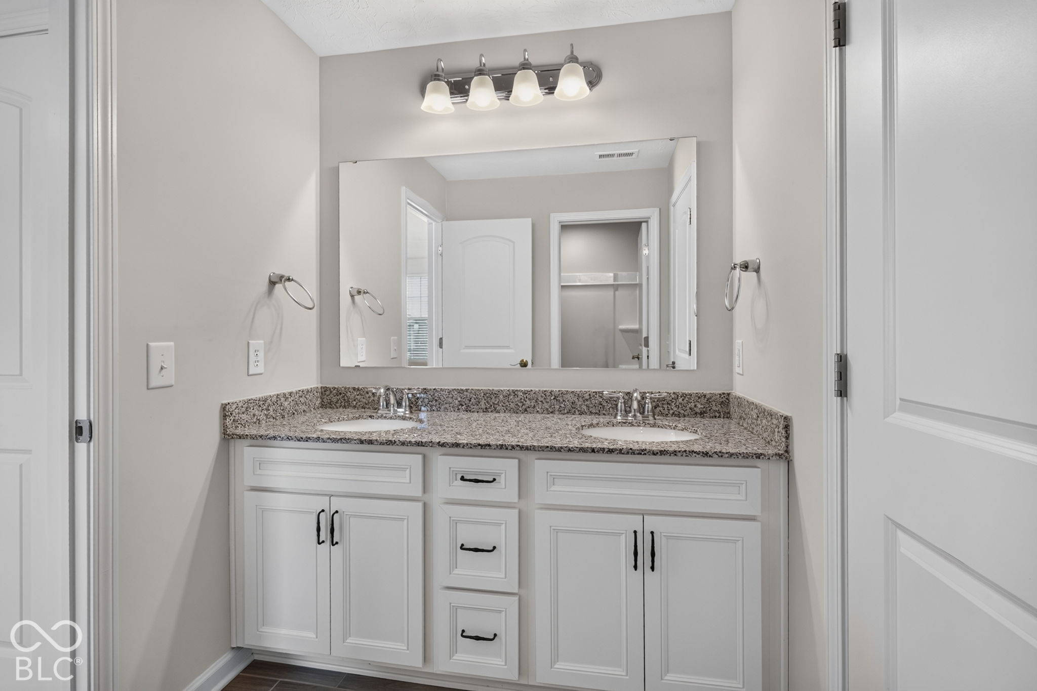 Double vanity featuring a stone countertop with dual undermount sinks, white cabinetry with matte black hardware, a full-width vanity mirror, and four-light overhead vanity fixture
