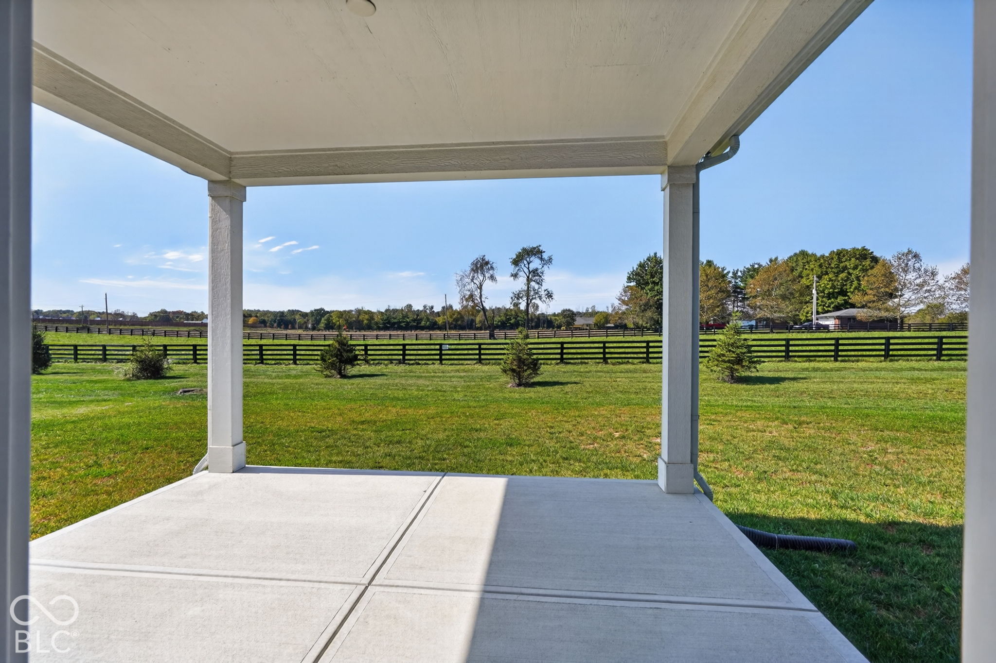 Covered concrete patio with structural columns overlooking an expansive lawn, split-rail fencing, and mature trees