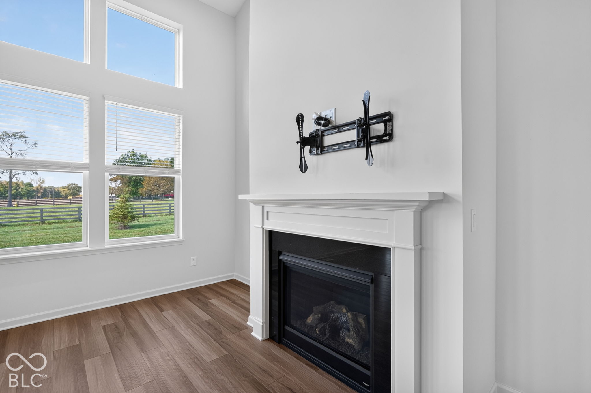 High-ceilinged room featuring wood-finish flooring, floor-to-ceiling windows, and a fireplace with a white mantel and black surround