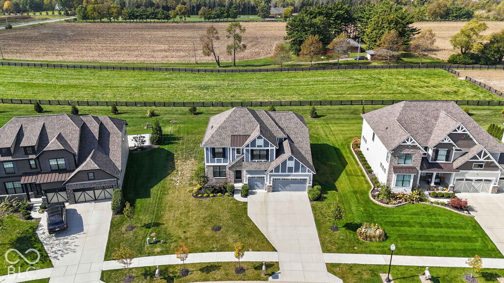 Aerial perspective of suburban residences featuring multi-gabled rooflines, stone and siding exteriors, attached multi-car garages, and concrete driveways, situated on expansive grounds with perimeter fencing