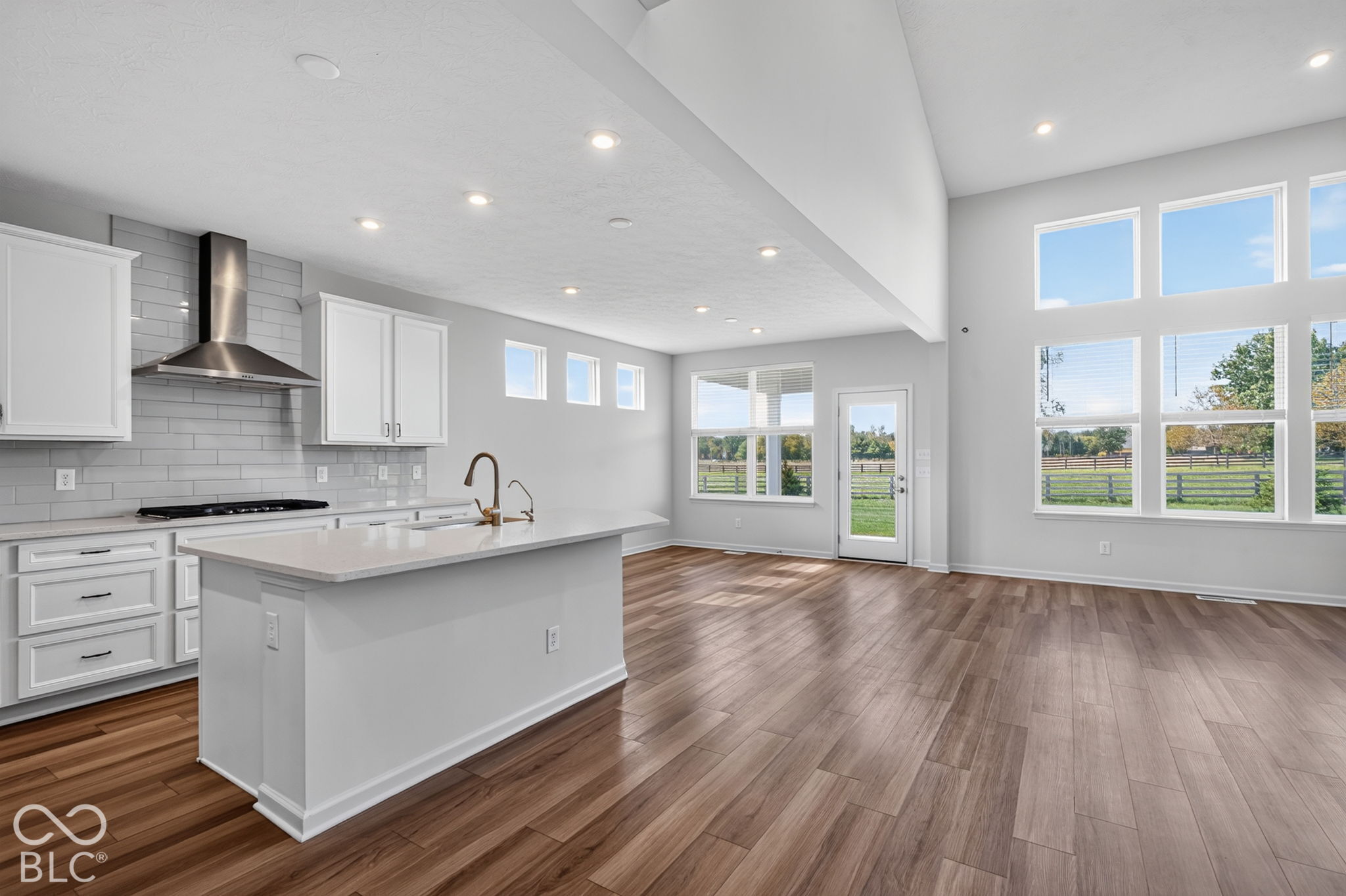 Open-concept kitchen featuring white cabinetry, a center island with stone-surface countertops, subway tile backsplash, stainless steel range hood, and wood-finish flooring under vaulted ceilings with expansive windows