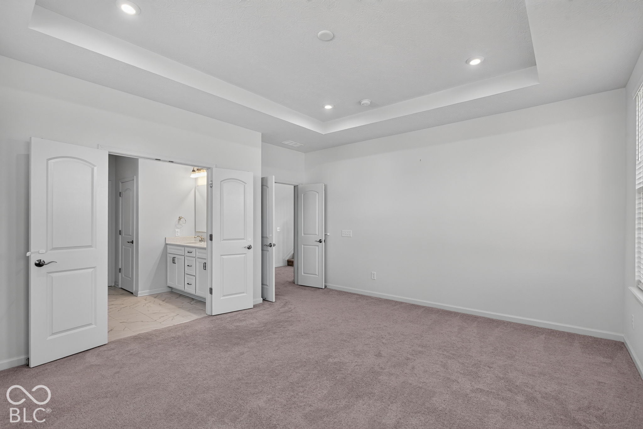 Tray ceiling with recessed lighting, wall-to-wall carpeting, and double doors opening to a vanity area with white cabinetry