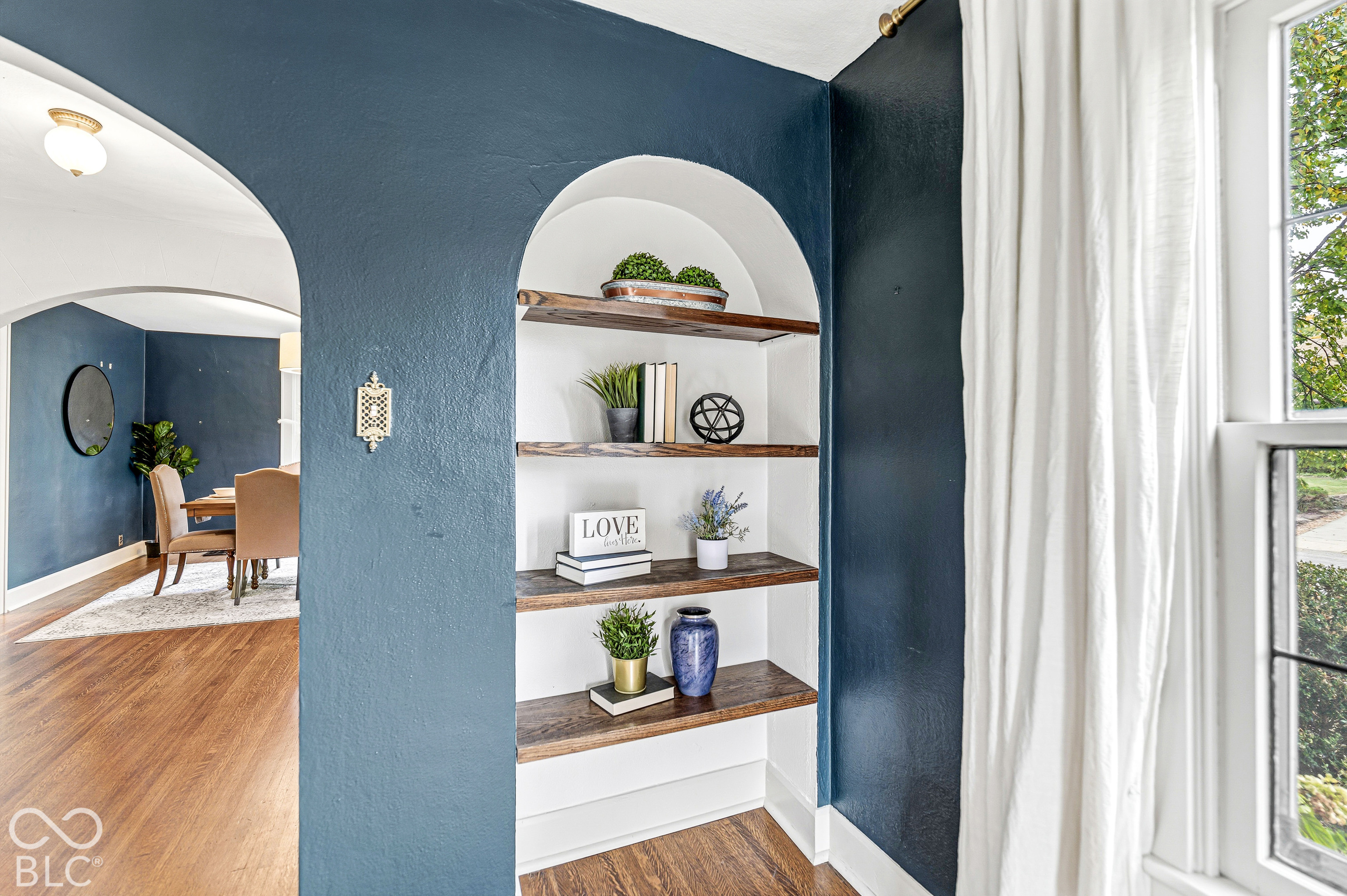 Arched built-in shelving with wood-finish floating shelves, expansive arched wall opening, wood-finish flooring, and multi-pane window