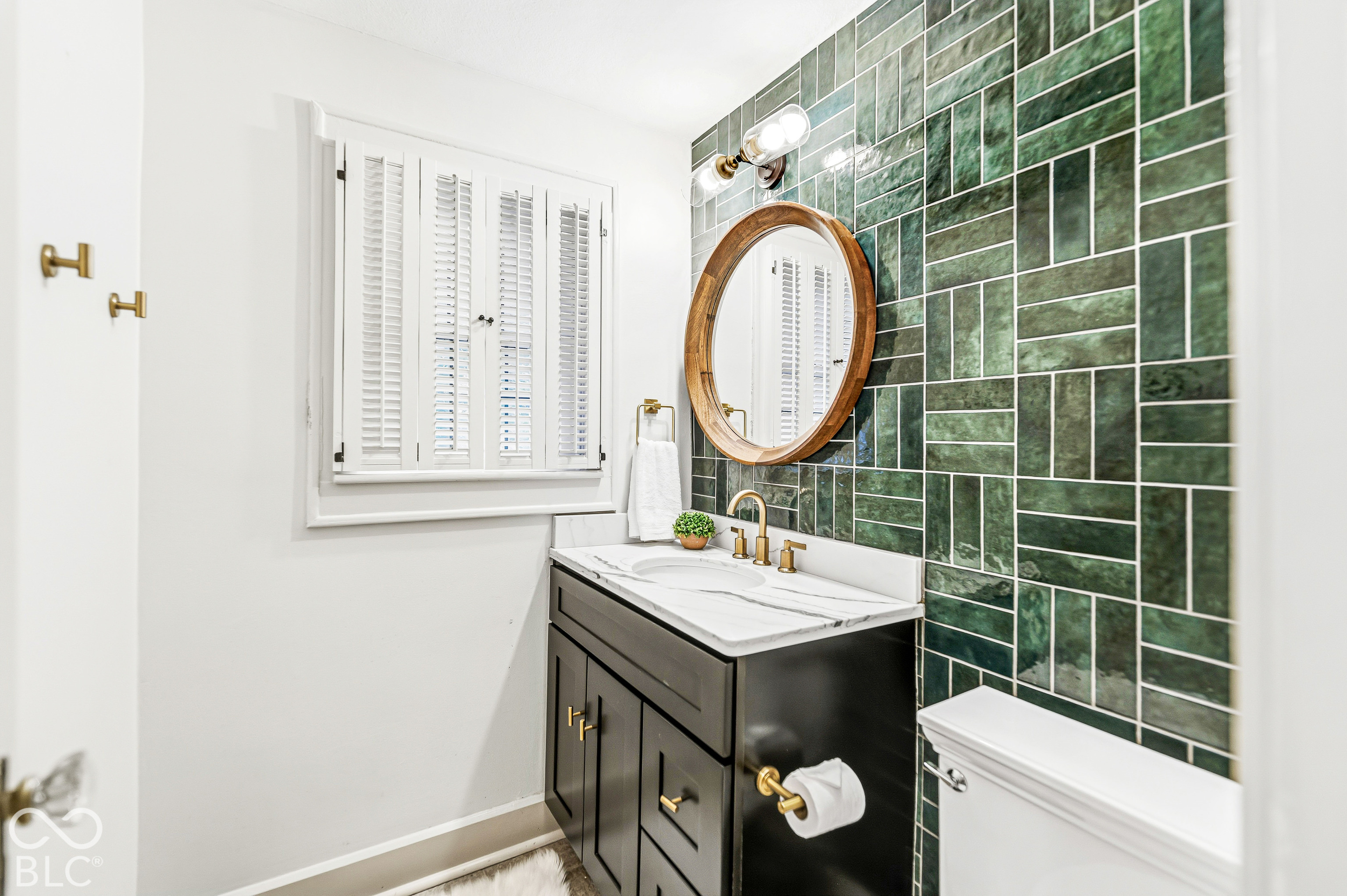 Bathroom featuring an emerald green tiled feature wall, a black vanity with a stone countertop, brass hardware, and white plantation shutters