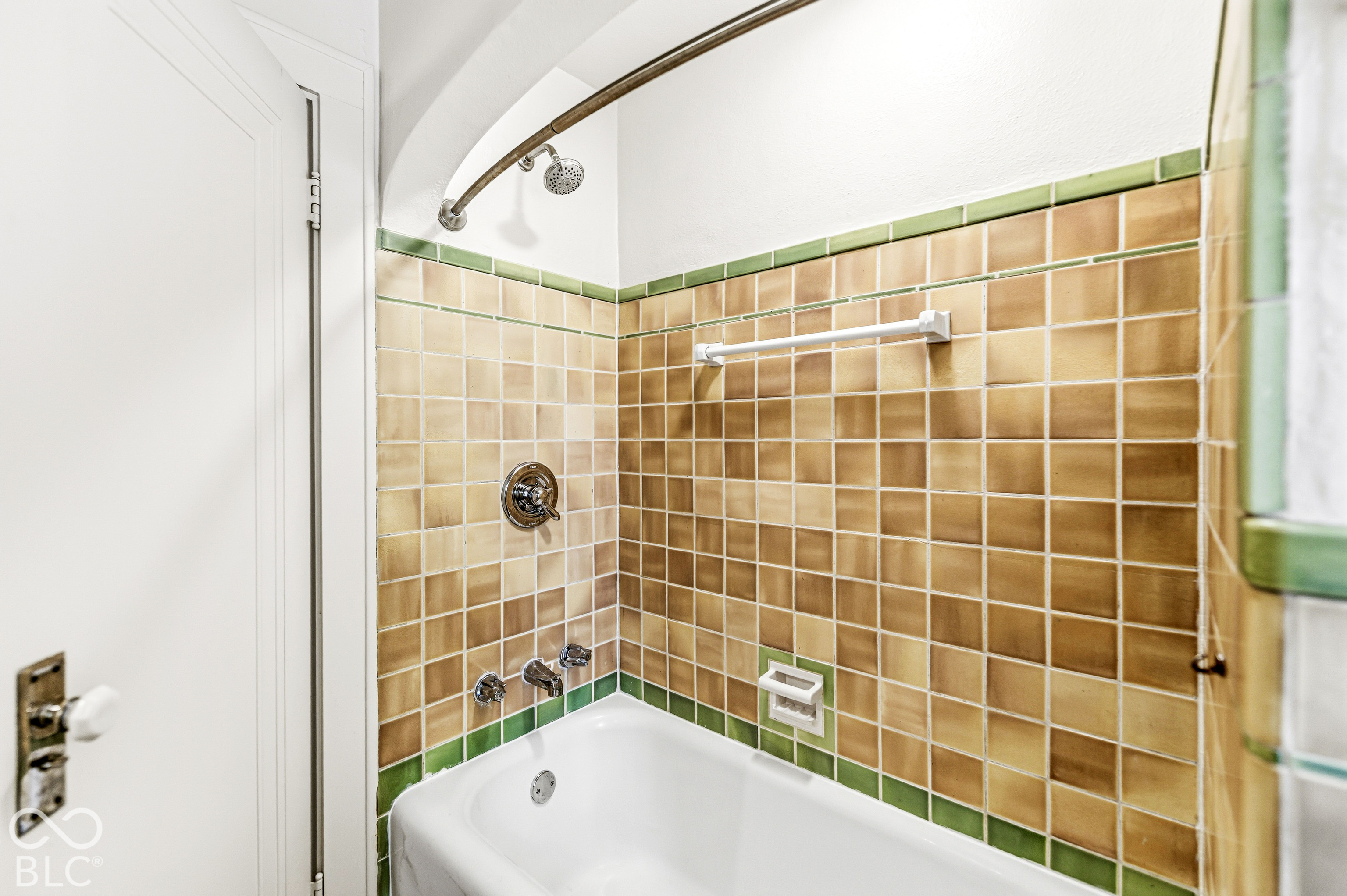 Bathtub surround featuring square ceramic wall tiling with decorative green trim, a curved shower rod, built-in soap dish, and wall-mounted metal plumbing fixtures