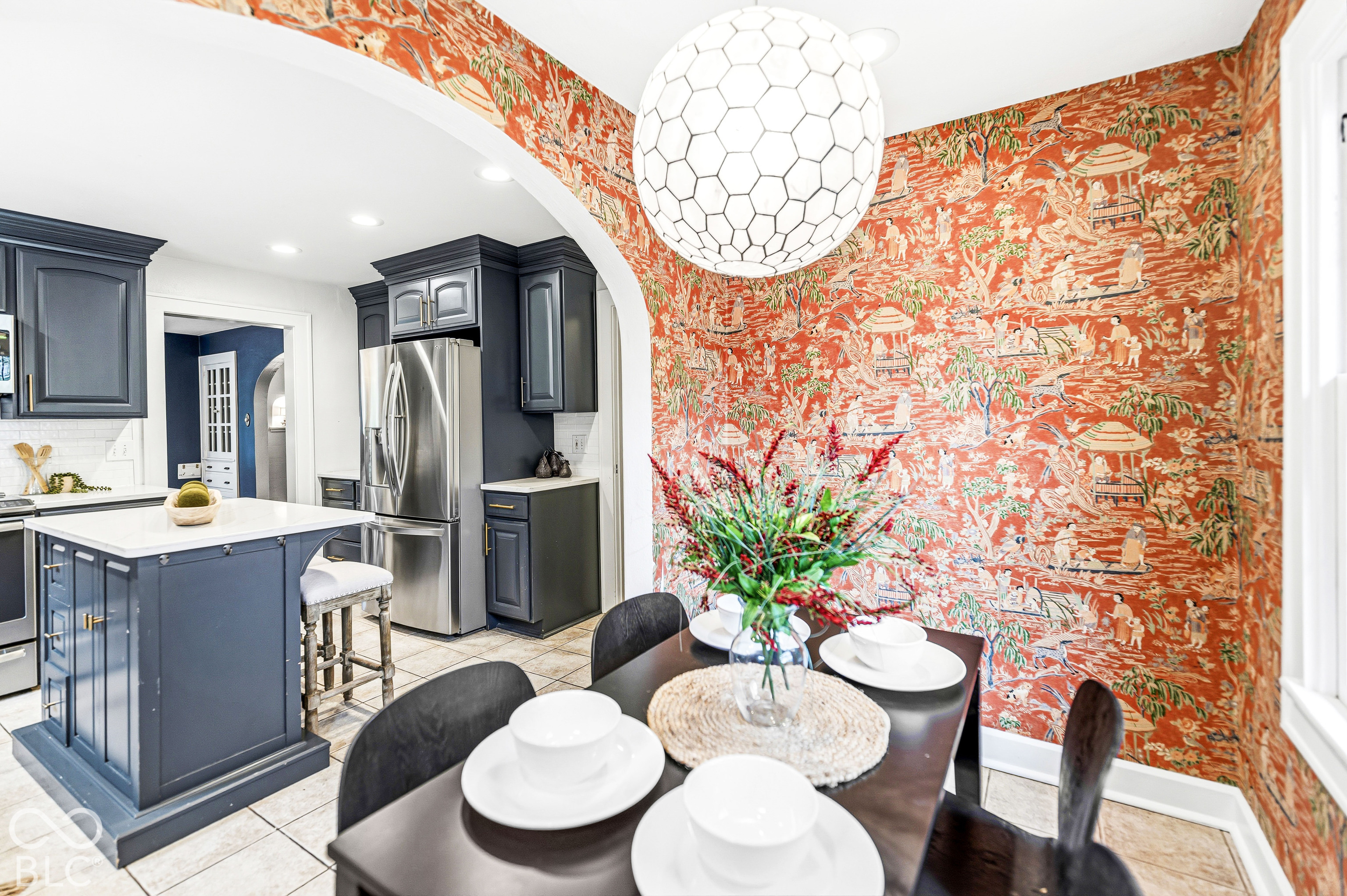 Kitchen featuring dark-toned cabinetry, a center island with a stone countertop, an arched architectural opening, patterned wall covering, and a modern geometric pendant light