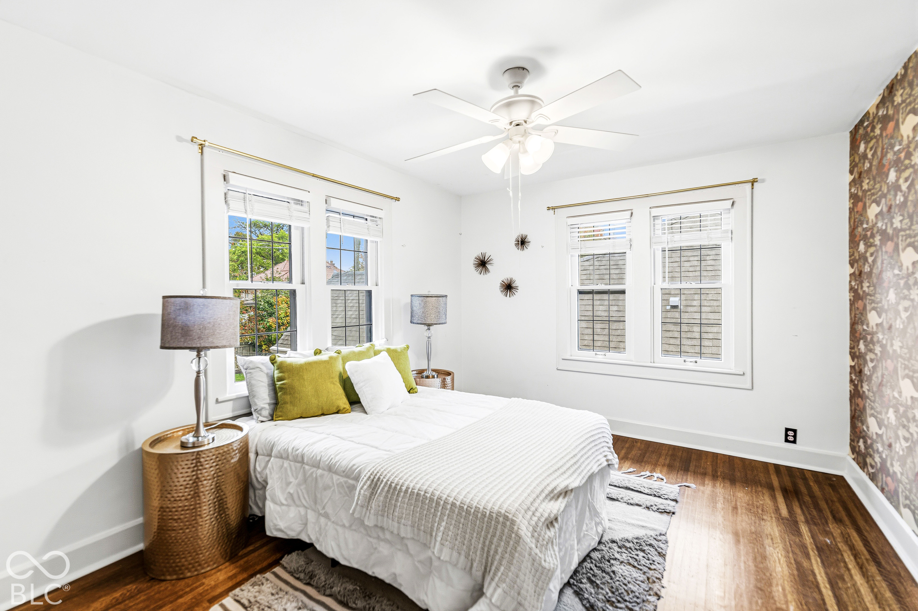 Room featuring wood-finish flooring, accent wallpaper, four windows with white trim, and a multi-blade ceiling fan with integrated lighting