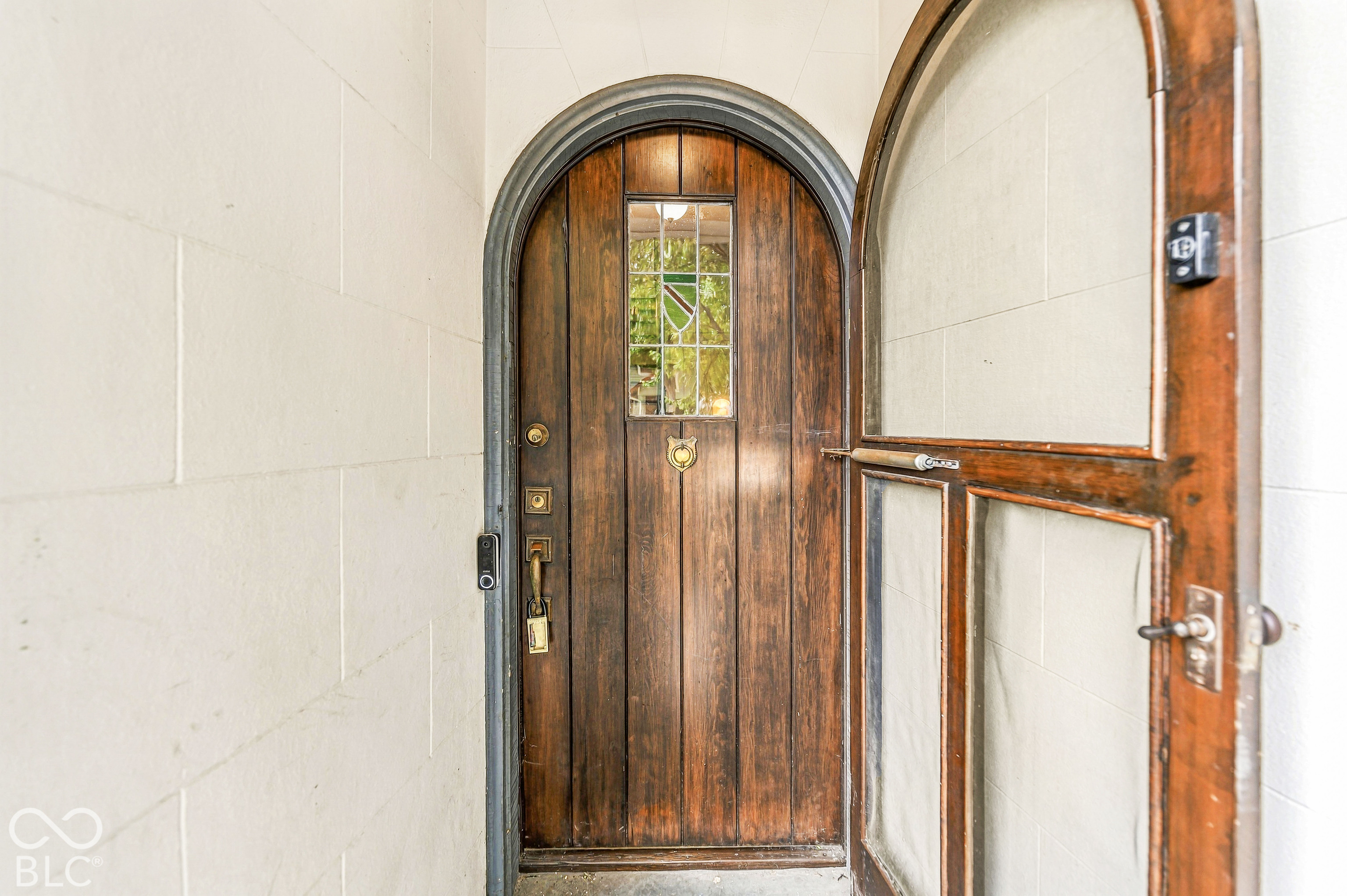 Arched solid wood front door featuring vertical plank construction, decorative leaded glass inlay, brass hardware, and a matching arched wood-frame storm door set within a light-toned masonry entryway