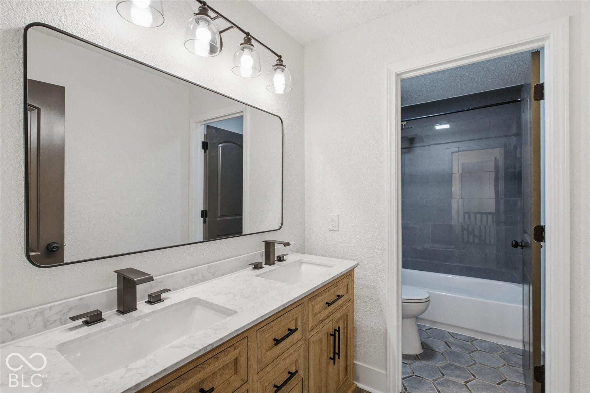 Dual-sink vanity with stone-finish countertop, dark metal fixtures, expansive rectangular mirror, and four-light vanity fixture