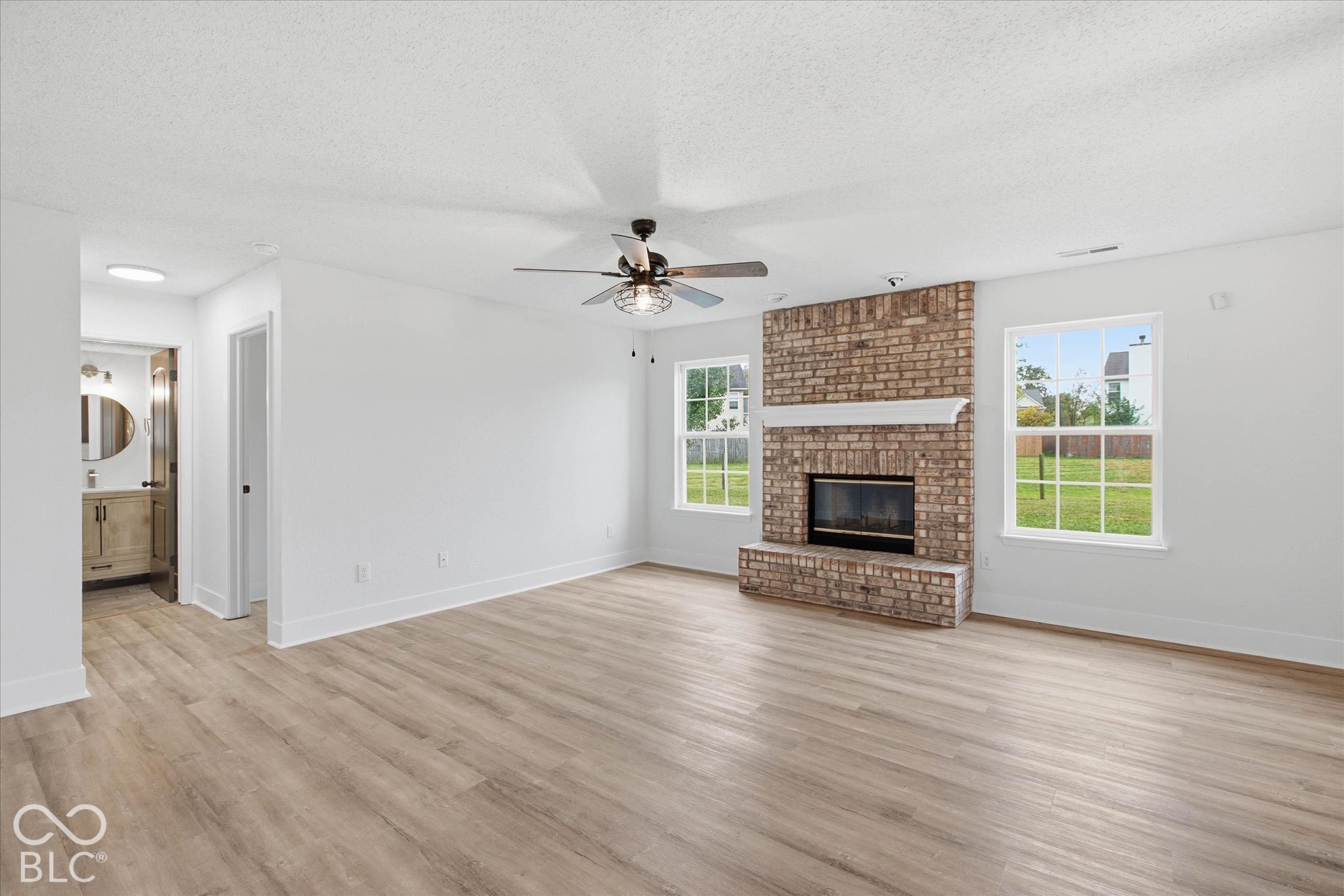 Living area featuring wood-finish flooring, floor-to-ceiling brick fireplace with a mantle, and a ceiling fan with integrated lighting
