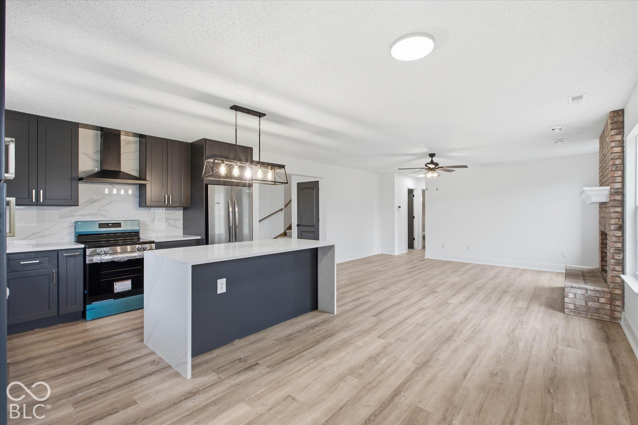 Kitchen featuring dark cabinetry, stone-style waterfall island, stainless steel range with overhead hood, wood-finish flooring, and a brick fireplace