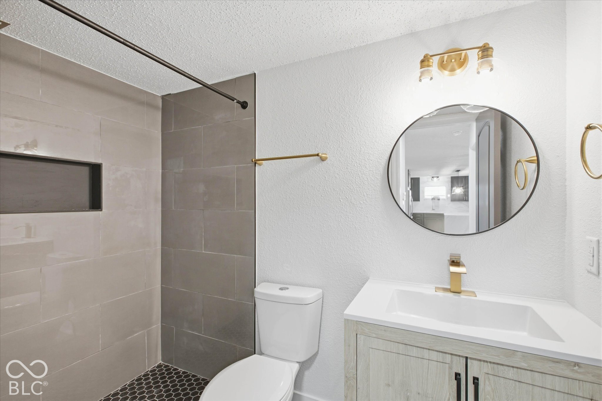 Bathroom featuring a wood-finish vanity with a white countertop, circular mirror, metallic wall-mounted lighting, and a shower enclosure with large-format wall tile, built-in shelving, and dark hexagonal floor tile
