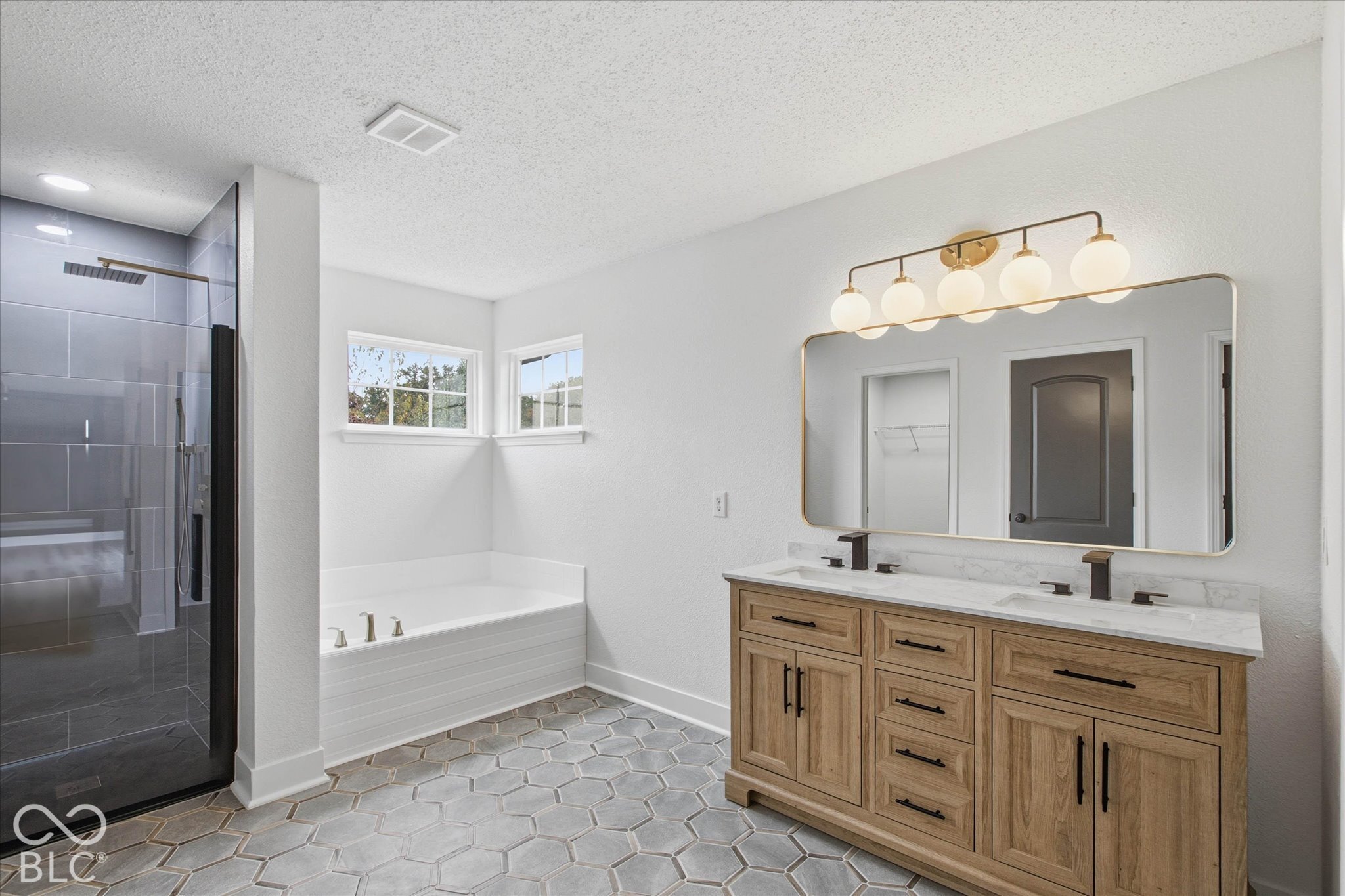Dual-sink vanity with light wood cabinetry, stone-finish countertop, and matte black fixtures, paired with hexagonal tile flooring, a soaking tub, and a glass-enclosed walk-in shower