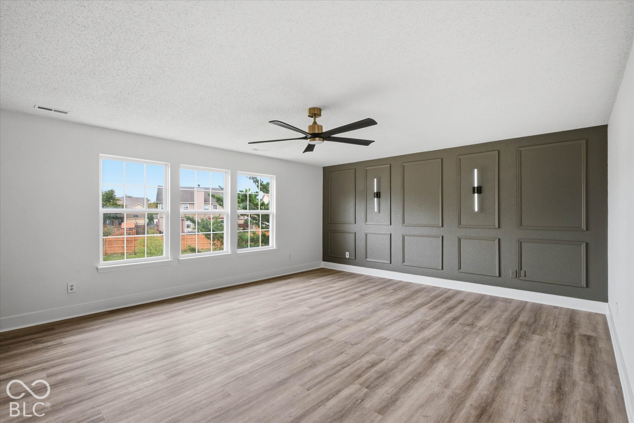 Room featuring wood-finish flooring, a charcoal-toned accent wall with decorative molding, dual wall-mounted light fixtures, a multi-blade ceiling fan, and large windows