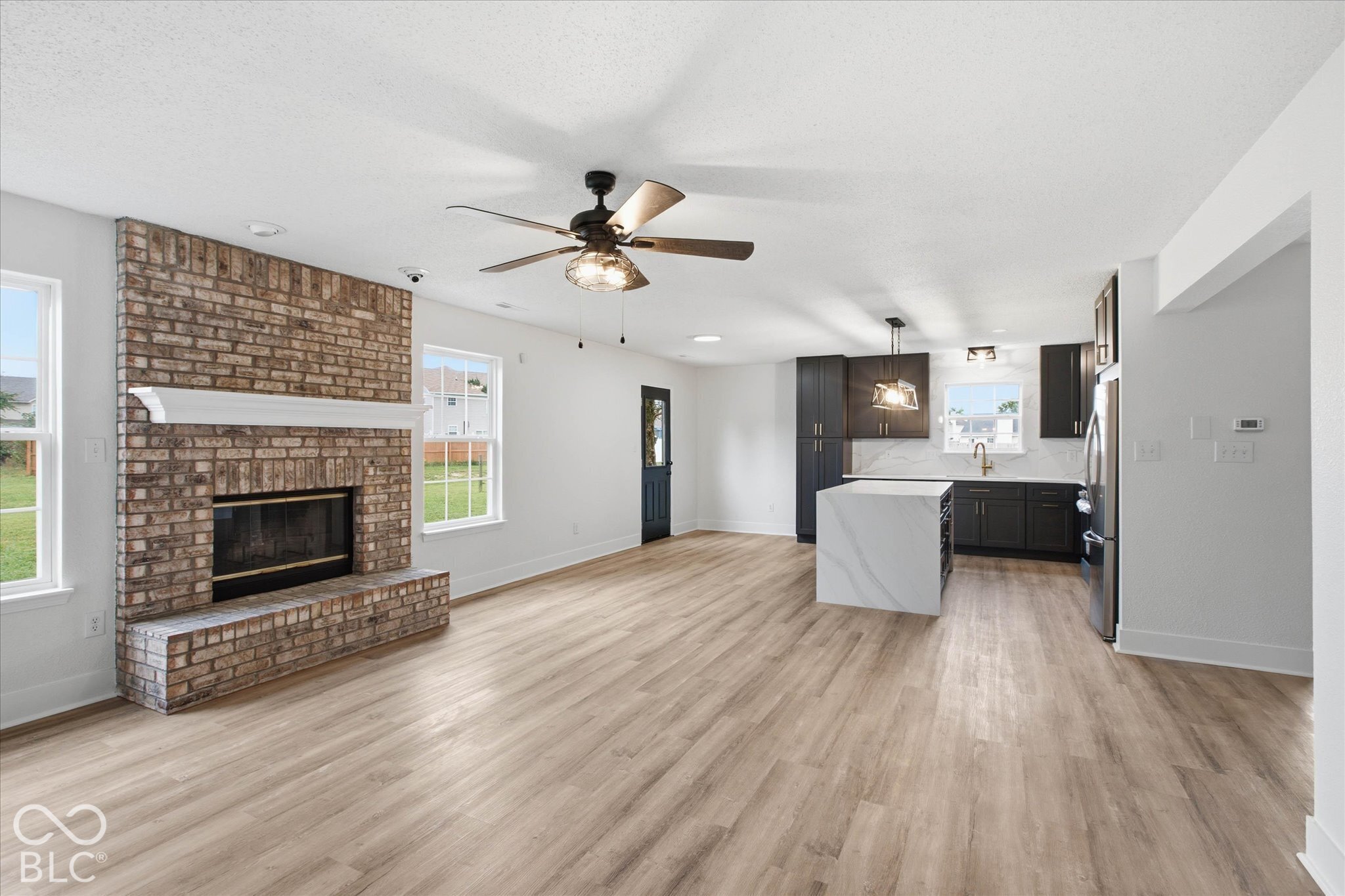 Full-height brick fireplace with a wood mantel, wood-finish flooring, kitchen island with a stone-finish waterfall countertop, dark-finish cabinetry, and stainless steel appliances