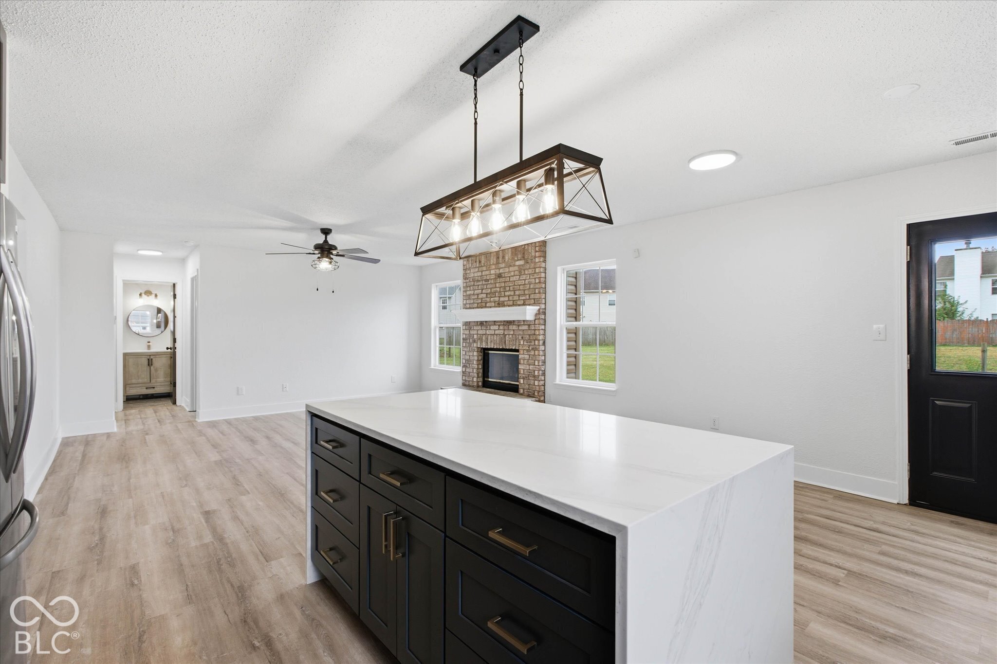 Kitchen island with solid-surface countertop and dark-finish cabinetry, wood-finish flooring, brick-accented fireplace, and industrial-style metal chandelier