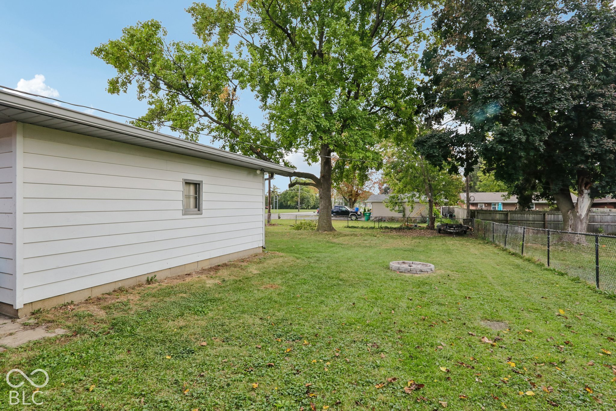 Backyard featuring horizontal vinyl siding, mature trees, chain-link fencing, and a stone-bordered fire pit