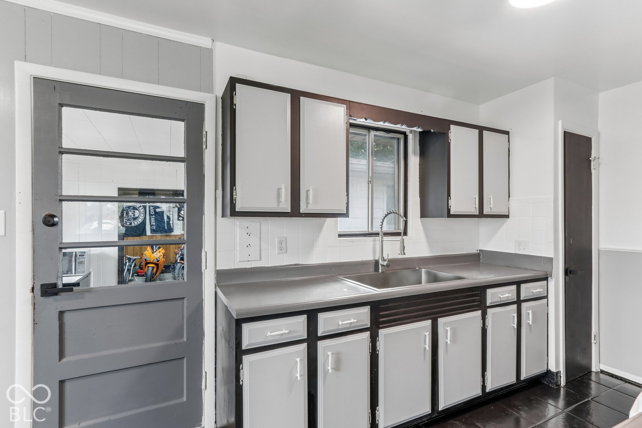 Kitchen featuring contrast-painted cabinetry, stainless steel sink with gooseneck faucet, solid-surface countertops, tile backsplash, and paneled exterior door with glass insert