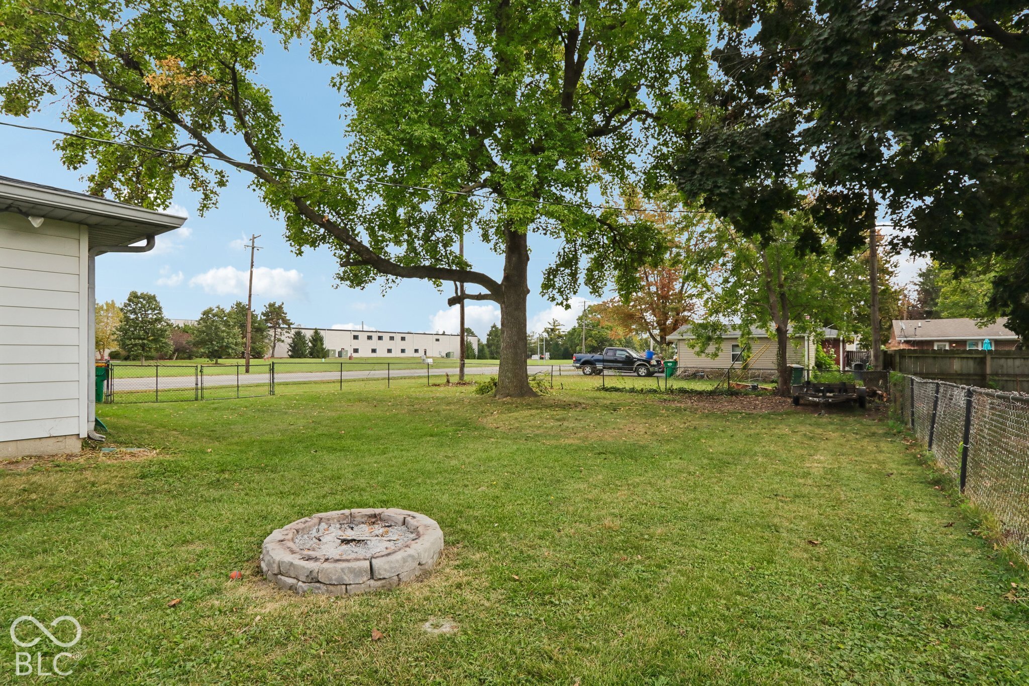 Expansive backyard featuring a stone fire pit, chain-link perimeter fencing, and a mature tree canopy