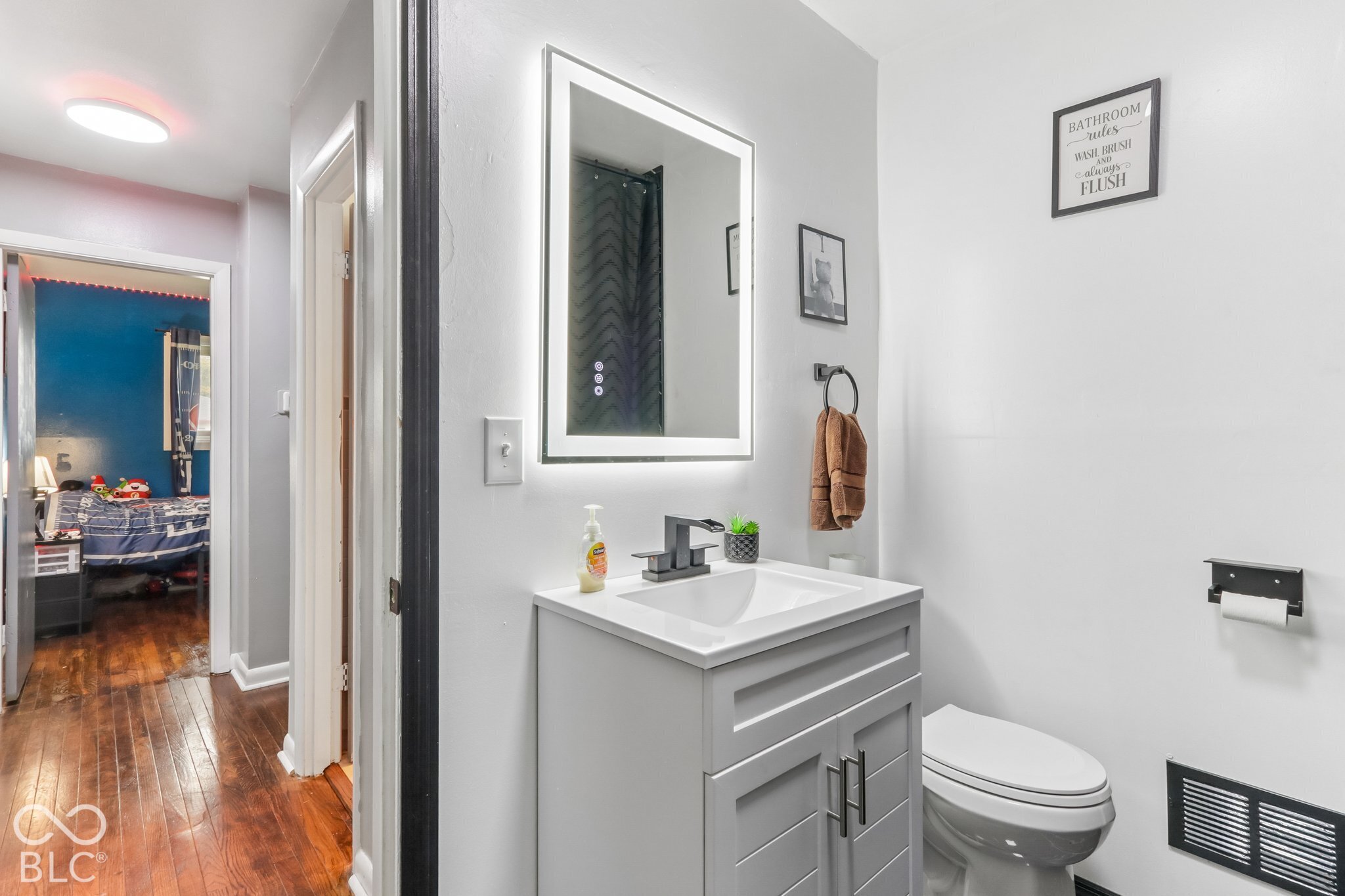 Bathroom featuring a grey vanity with a white integrated countertop, matte black faucet, LED-backlit mirror, and matte black hardware