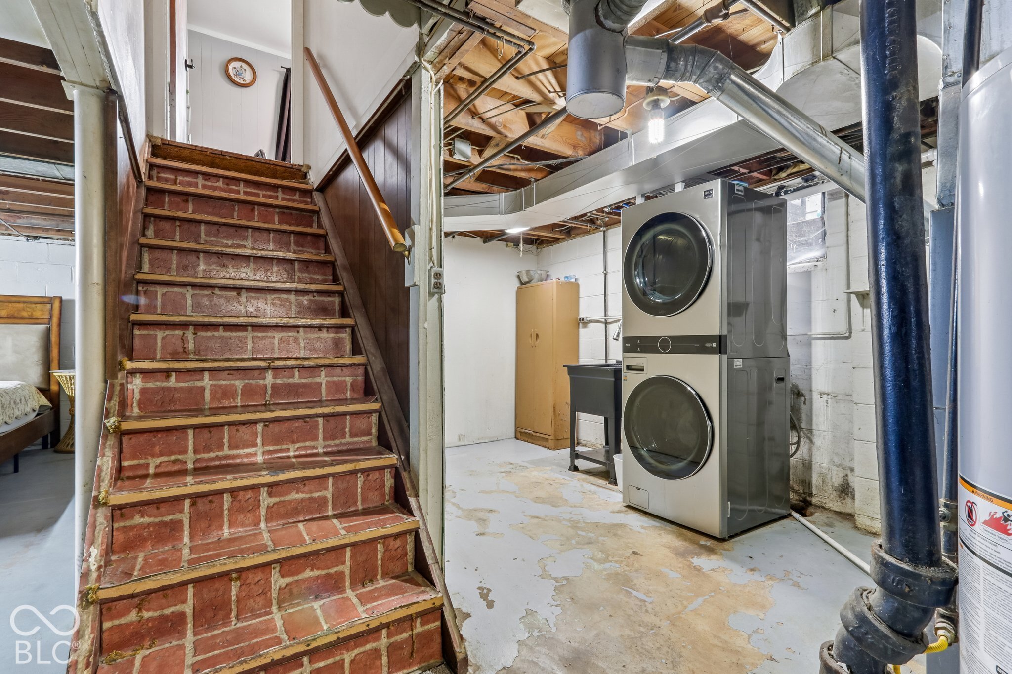 Lower-level utility area featuring a stacked laundry unit, brick-clad staircase with a wooden handrail, utility sink, and painted concrete flooring