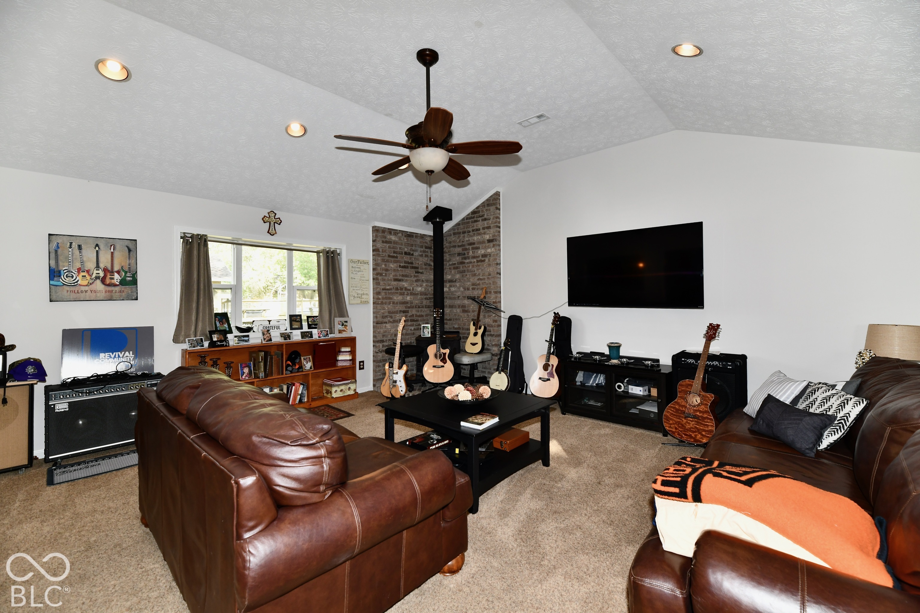 Vaulted ceiling featuring recessed lighting and a central ceiling fan, highlighted by a floor-to-ceiling brick accent wall with a wood-burning stove and neutral wall-to-wall carpeting