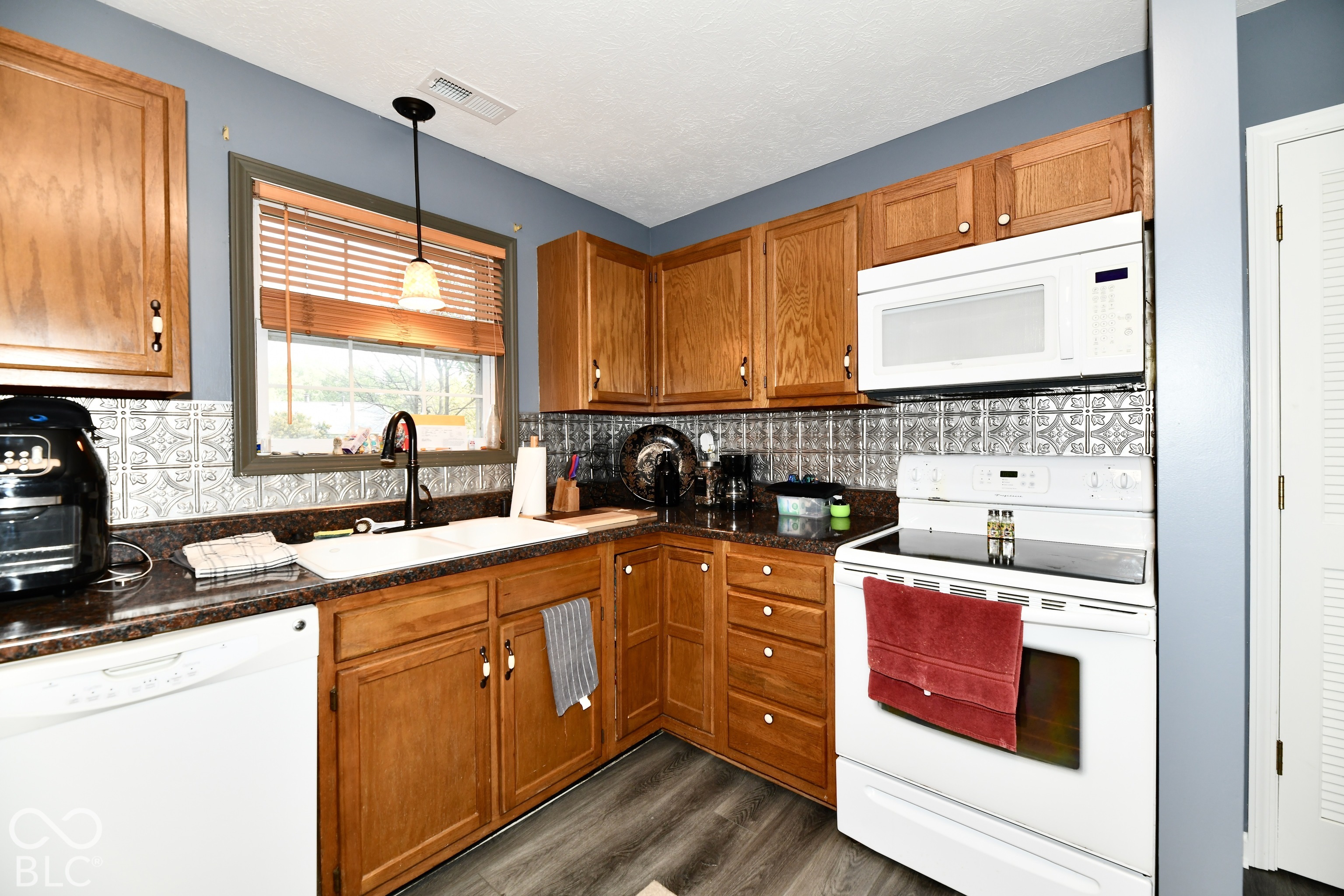 Kitchen featuring wood cabinetry, patterned tile backsplash, dark solid-surface countertops, dual-basin white sink with a matte black faucet, and wood-finish flooring