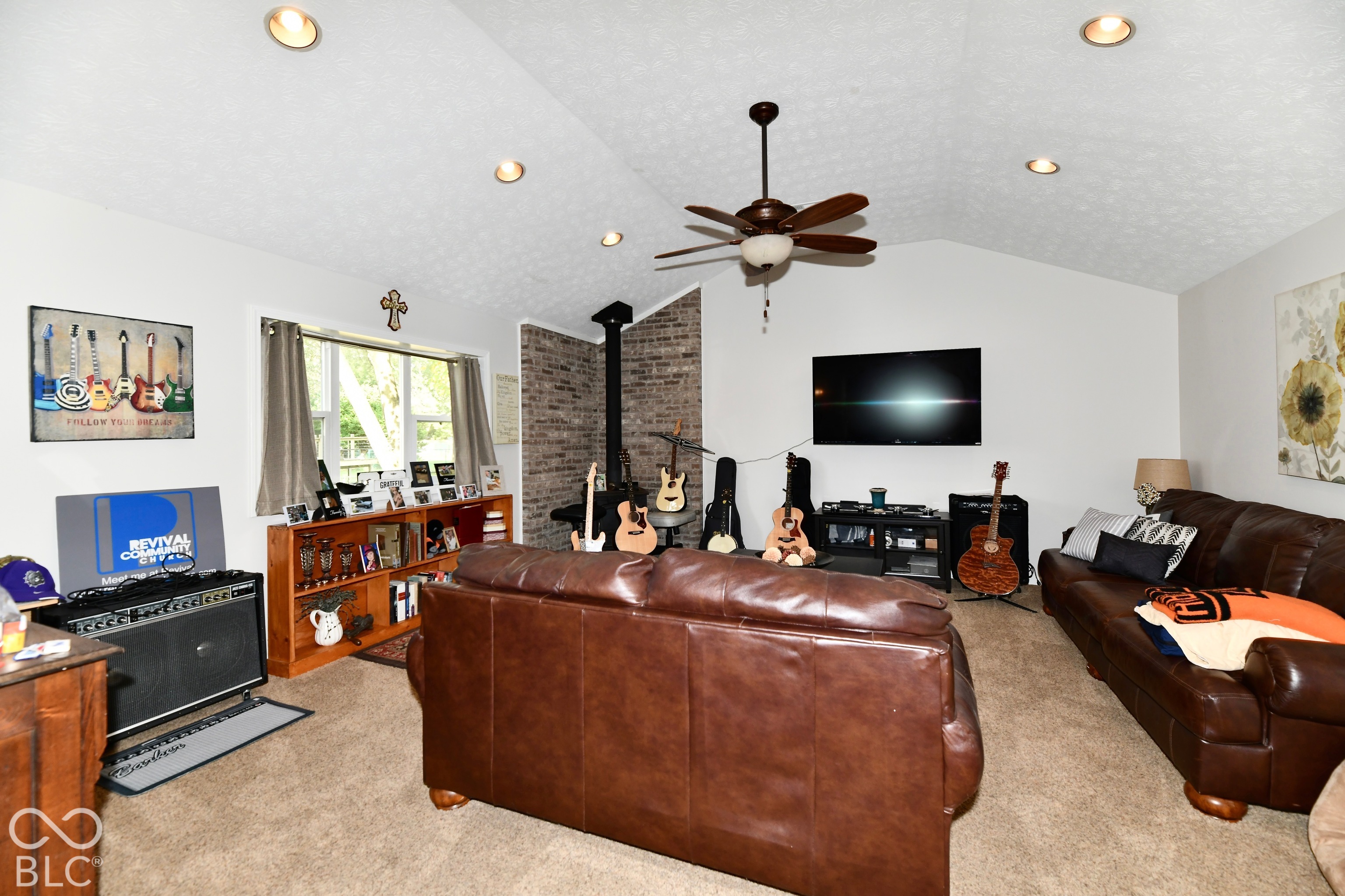 Vaulted ceiling featuring recessed lighting and a central ceiling fan, exposed brick accent wall with a wood-burning stove, and a window with exterior light exposure